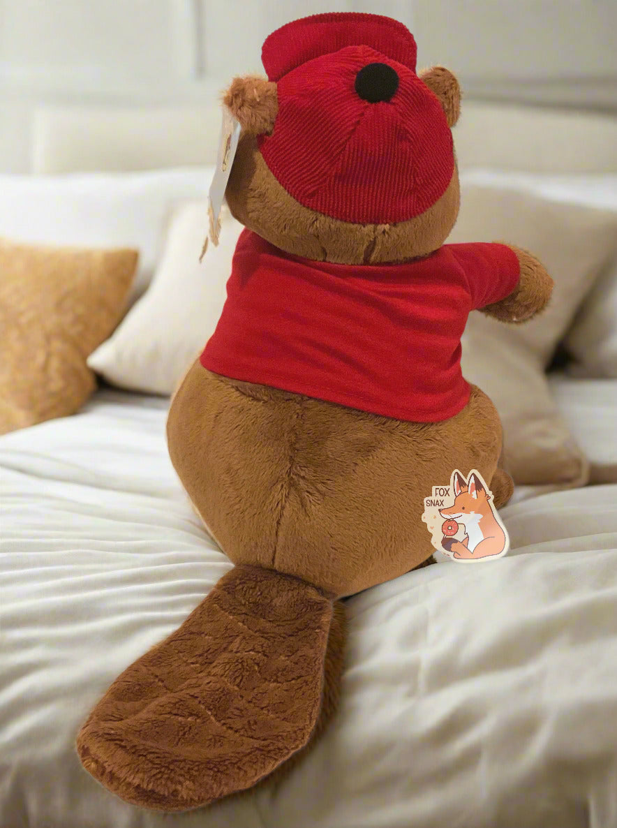 A picture of the back of an adorable Buc-ee the Beaver plush. The plush is of Buc-ee, wearing a red Buc-ee's shirt and red ball cap, and is looking to the left. Buc-ee has a large brown fuzzy beaver tail, and is covered in plush brown and white stuffed animal "fur". The background is of a fluffy warm bed in a bedroom.