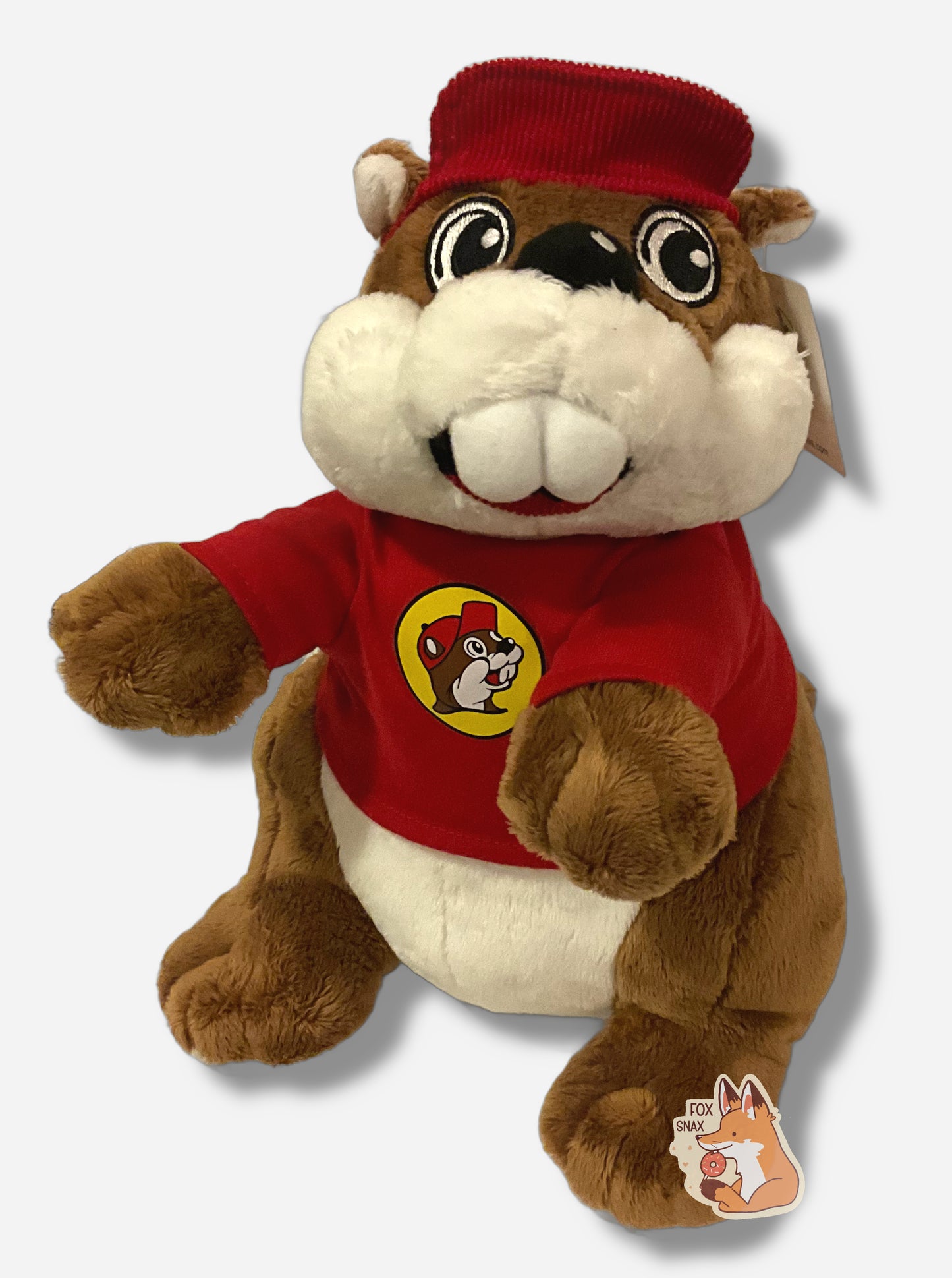 A picture of an adorable Buc-ee the Beaver plush. The plush is of Buc-ee, wearing a red Buc-ee's shirt and red ball cap, and is looking to the left. It is roughly a foot tall and covered in plush brown and white stuffed animal "fur".