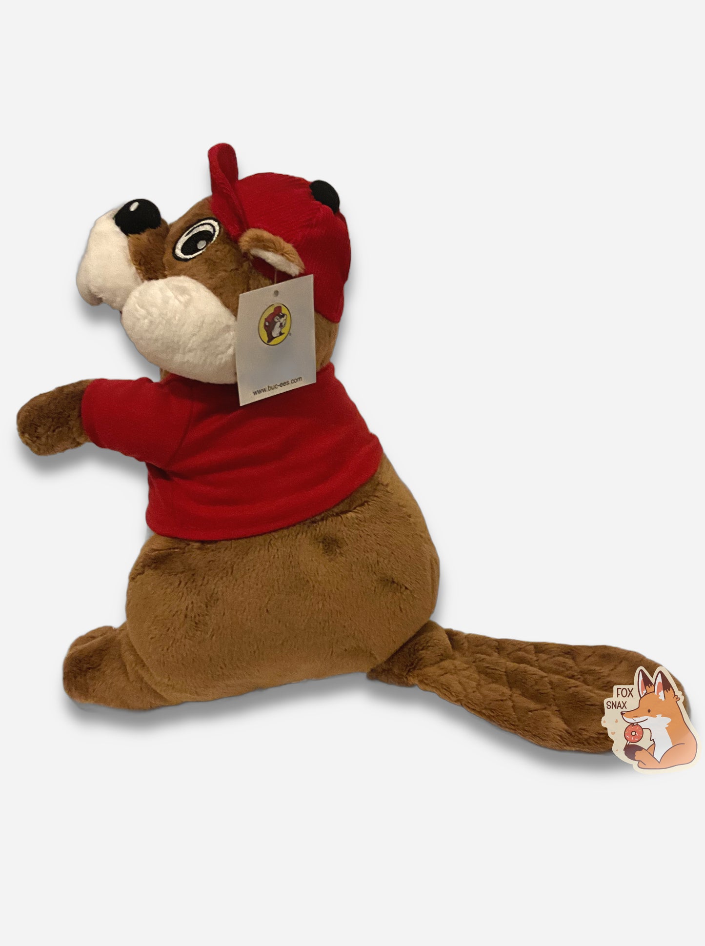 A picture of the side of an adorable Buc-ee the Beaver plush. The plush is of Buc-ee, wearing a red Buc-ee's shirt and red ball cap, and is looking to the left. Buc-ee has a large brown fuzzy beaver tail, and is covered in plush brown and white stuffed animal "fur".