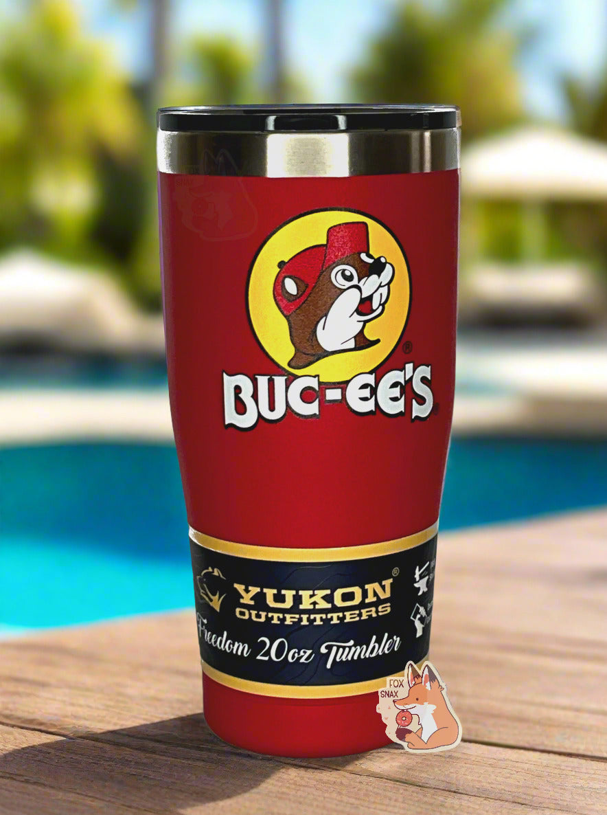A picture of a tall, red, tumbler, with a big picture of Buc-ee The Beaver on it. The surface is smooth and red, as it is red paint covering stainless steel. There is no handle. The top is plastic. The top of the mug is lined with a stainless steel band. A paper wrapper around the bottom reads: Yukon Outfitters. Freedom 20 oz Tumbler.