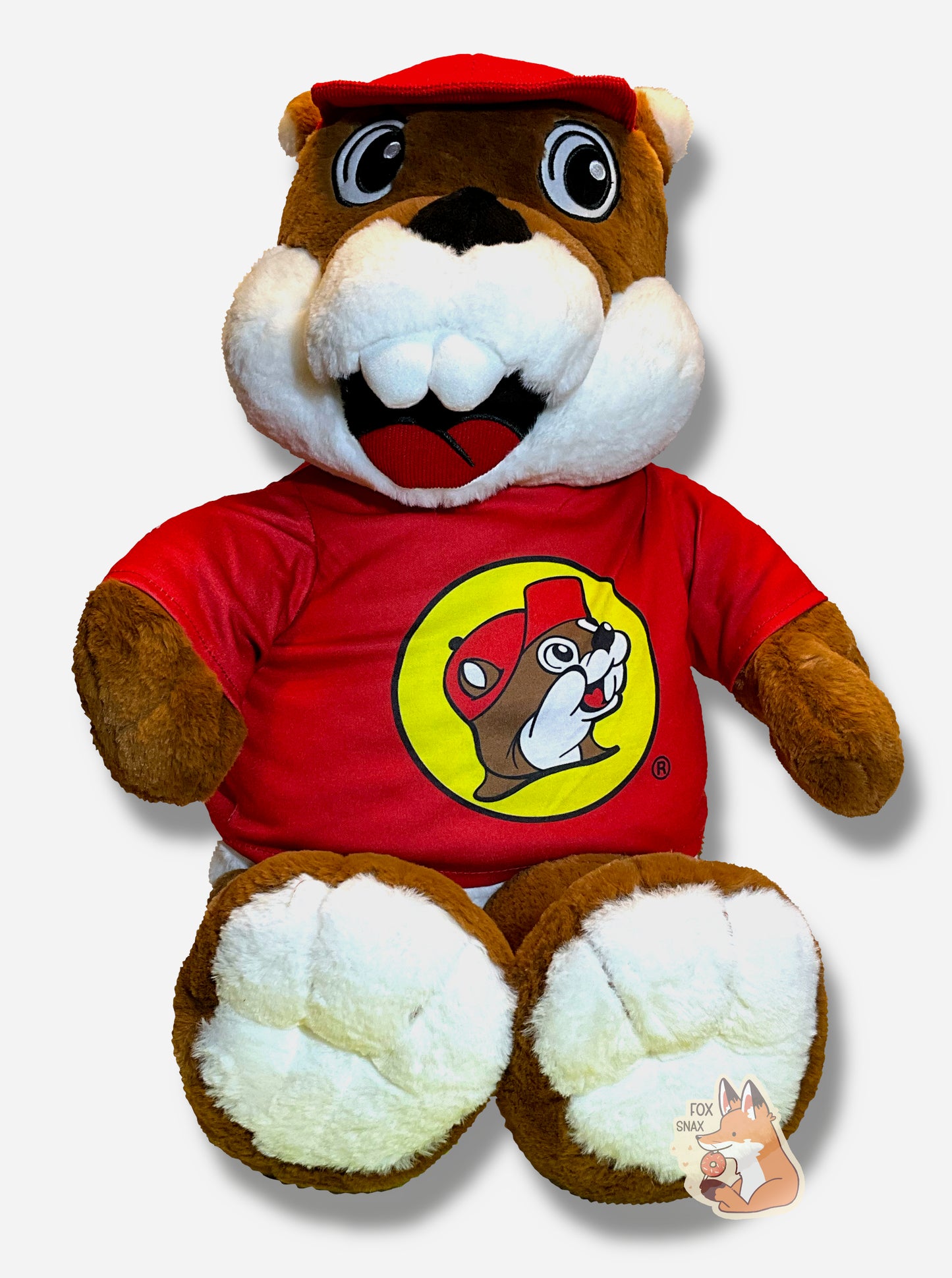 A picture of an adorable Buc-ee the Beaver plush. The plush is of Buc-ee, wearing a red Buc-ee's shirt and red ball cap, and is looking to the left. It is roughly two feet tall and covered in plush brown and white stuffed animal "fur".
