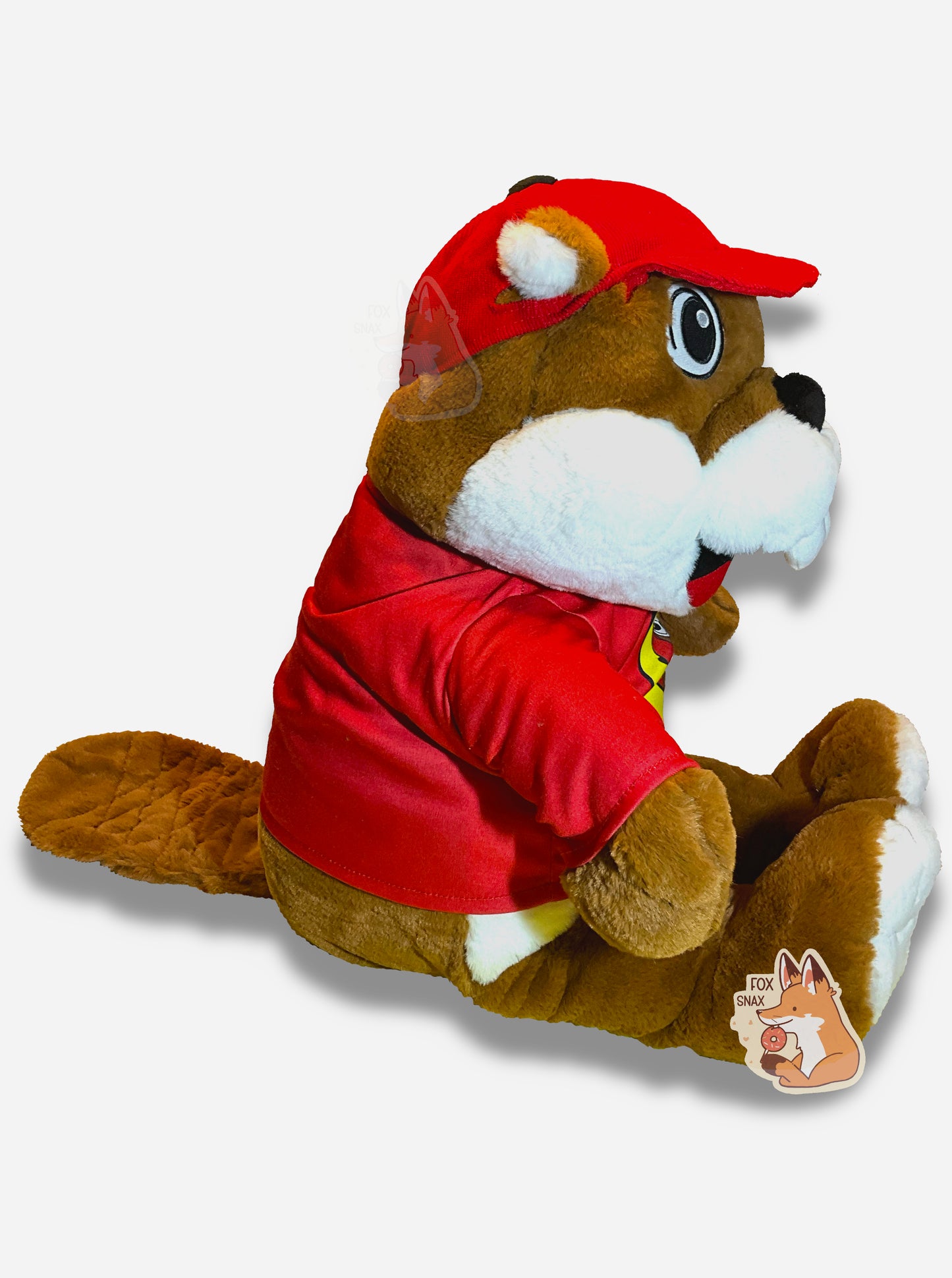 A picture of the side of an adorable Buc-ee the Beaver plush. The plush is of Buc-ee, wearing a red Buc-ee's shirt and red ball cap, and is looking to the left. Buc-ee has a large brown fuzzy beaver tail, and is covered in plush brown and white stuffed animal "fur".