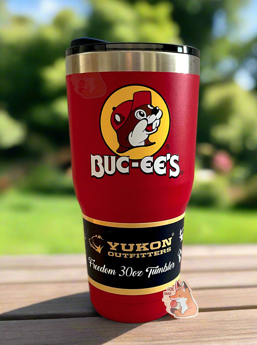A picture of a tall, red, tumbler, with a big picture of Buc-ee The Beaver on it. The surface is smooth and red, as it is red paint covering stainless steel. There is no handle. The top is plastic. The top of the mug is lined with a stainless steel band. A paper wrapper around the bottom reads: Yukon Outfitters. Freedom 30 oz Tumbler. The tumbler is sitting on a patio table against a pretty, manicured backyard lawn.