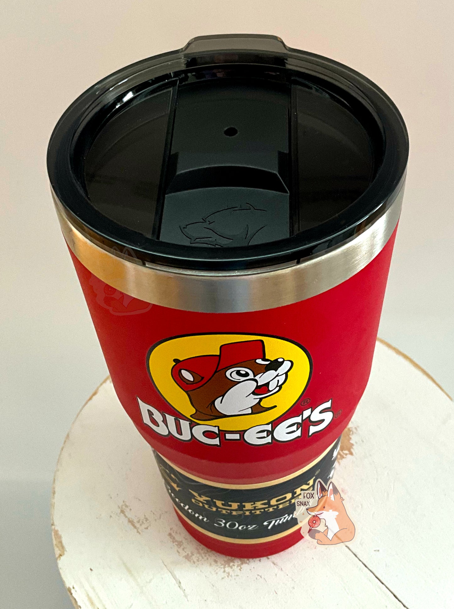 A three-quarters view of a picture of a tall, red, tumbler, with a big picture of Buc-ee The Beaver on it. The surface is smooth and red, as it is red paint covering stainless steel. There is no handle. The top is black plastic, and shows a slider in the middle with the imprint of the Yukon bear's head on it. The top of the mug is lined with a stainless steel band. A paper wrapper around the bottom reads: Yukon Outfitters. Freedom 30 oz Tumbler. The tumbler is sitting on a white circular stool.