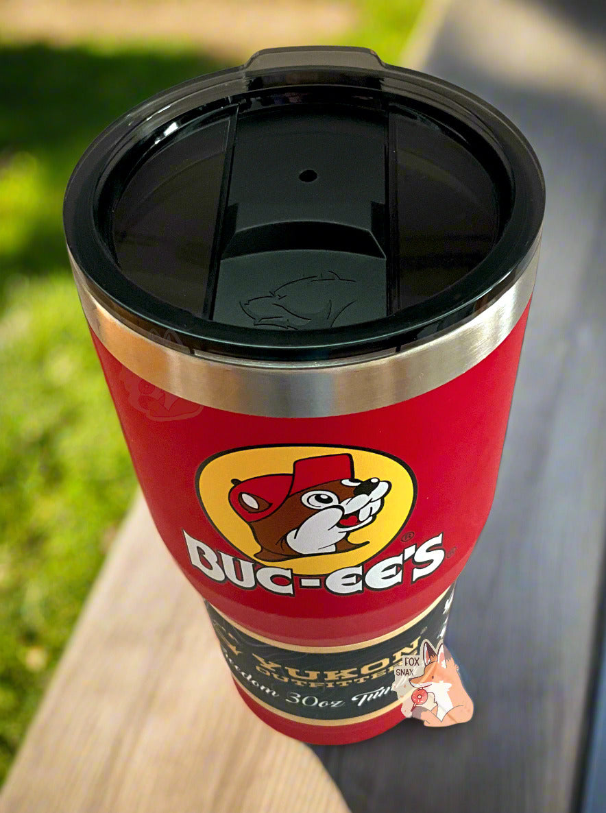 A three-quarters view of a picture of a tall, red, tumbler, with a big picture of Buc-ee The Beaver on it. The surface is smooth and red, as it is red paint covering stainless steel. There is no handle. The top is black plastic, and shows a slider in the middle with the imprint of the Yukon bear's head on it. The top of the mug is lined with a stainless steel band. A paper wrapper around the bottom reads:Yukon Outfitters.The tumbler is sitting on a patio table against a pretty, manicured backyard lawn.