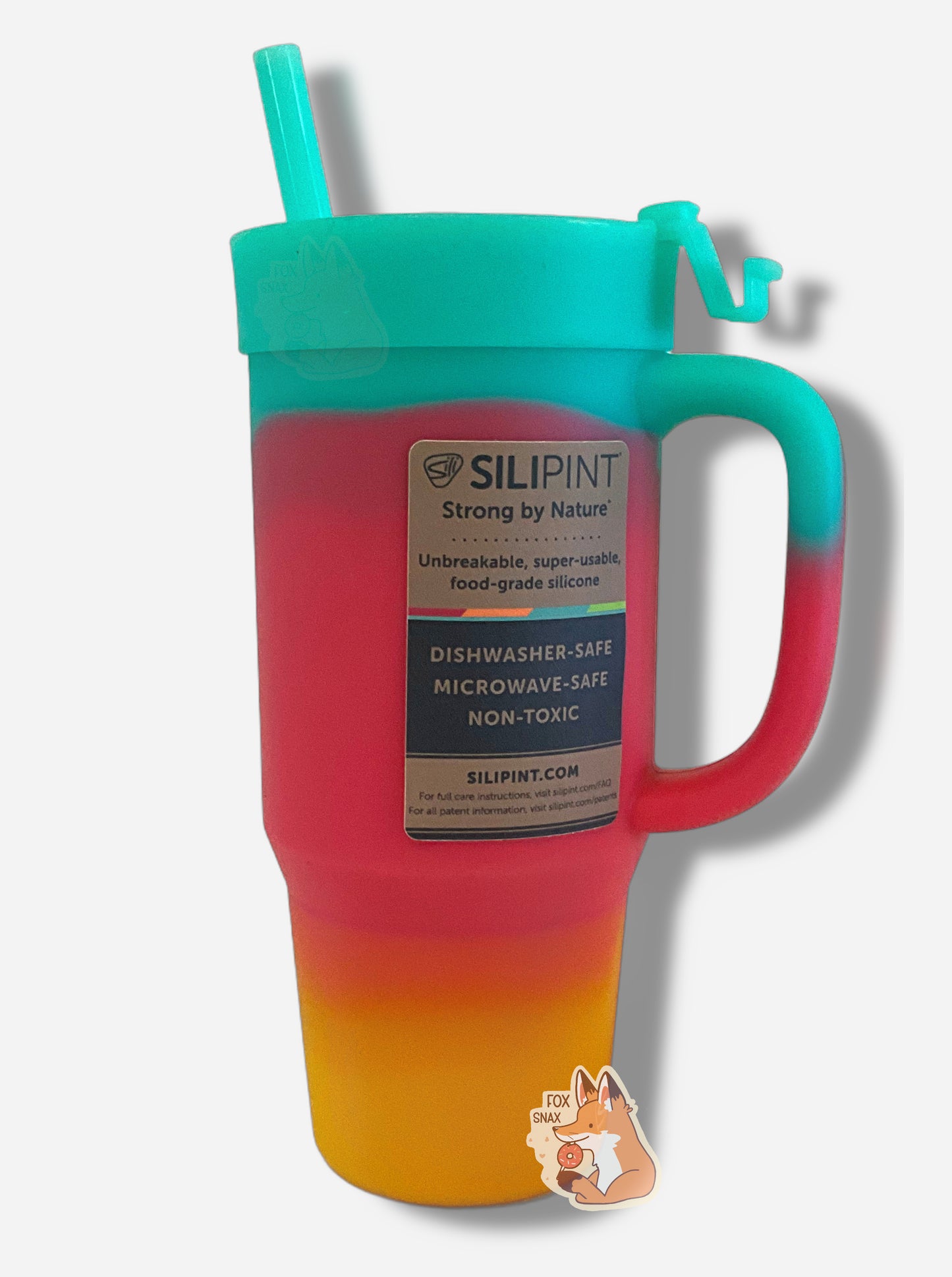 A picture of the back of a tall, rainbow colored tumbler. The surface starts as orange-yellow on the bottom, fading to red in the middle, then blue/green at the top. The entire mug is made of silicone, including the top and the straw. A brown sticker, in black font on the back, reads "SILIPINT, Strong By Nature, Unbreakable, super-usable, food-grade silicone. Dishwasher-safe, Microwave-safe, Non-toxic. Silipint.com"