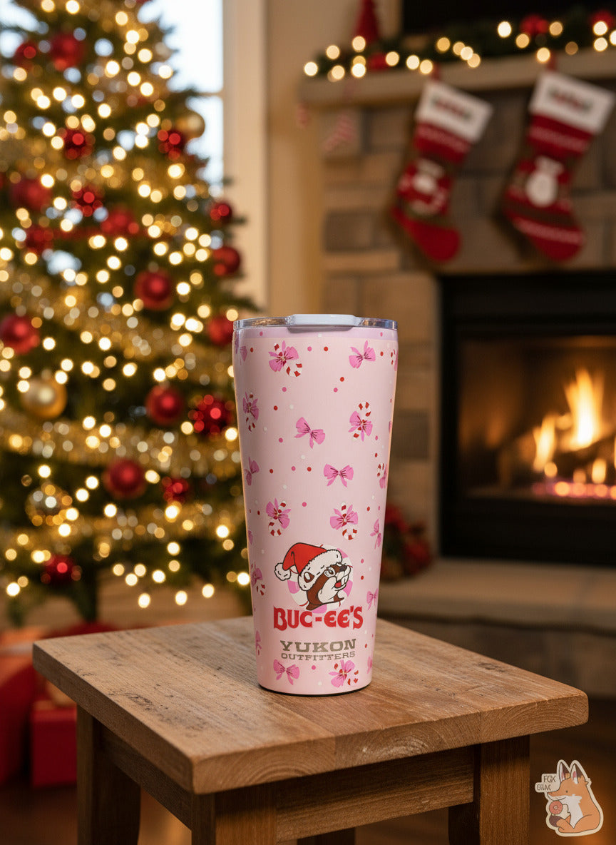 A picture of a pink double pint tumbler, with a light pink background full of bows and candy canes. The middle bottom of the tumbler has the Buc-ee's logo, but with Buc-ee The Beaver wearing a Santa hat, framed against a peppermint. The label on the bottom reads BUC-EE'S in red text, then underneath that, YUKON OUTFITTERS. The tumbler is sitting on a wooden footstool against a beautiful Christmas tree background, complete with fireplace and stockings on the mantel.