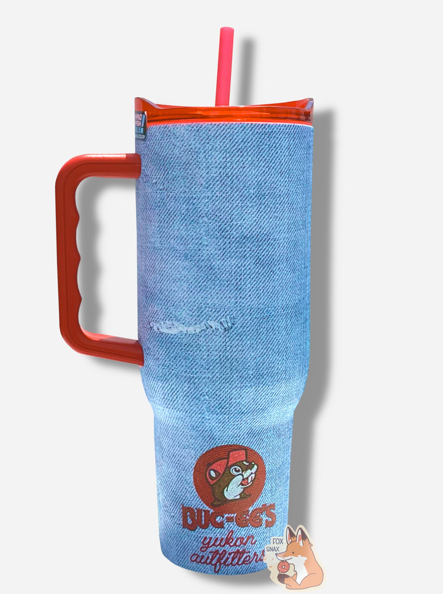 A picture of the back of a tall, faded denim blue, sorta skinny tumbler, with a small picture of Buc-ee The Beaver on it in the middle bottom. The surface is bumpy and light blue, as if made out of denim (it's not; it's metal). Several "rips" mar the surface; they show the white stitching underneath. The tumbler is light blue paint covering stainless steel. The handle, top, and straw are red plastic. The bottom icon reads: BUC-EE'S in red "stitching" letters, followed by "yukon outfitters" in cursive