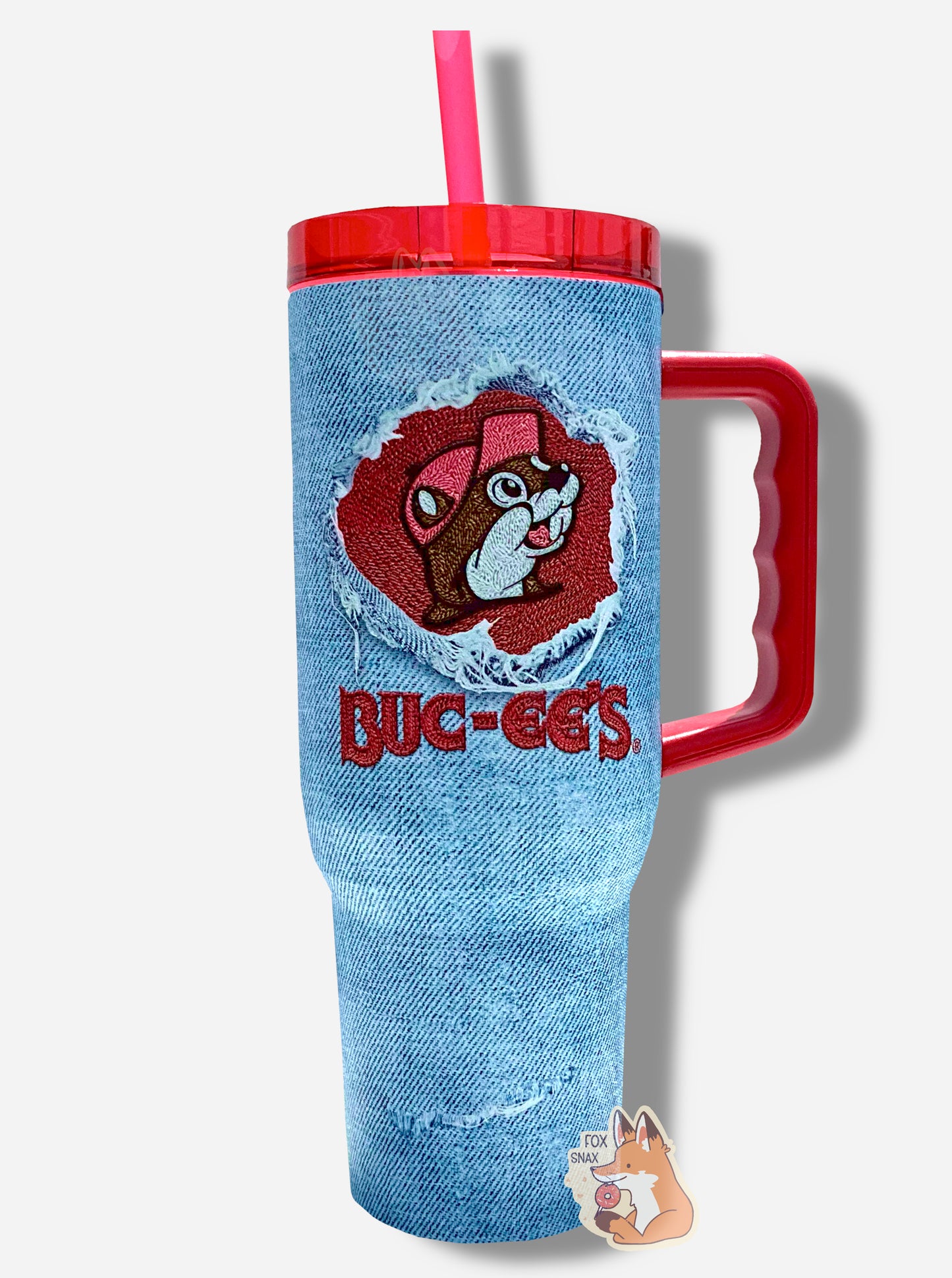 A picture of a tall, faded denim blue, sorta skinny tumbler, with a big picture of Buc-ee The Beaver on it in the middle, inside of a circular denim "rip". The surface is bumpy and light blue, as if made out of denim (it's not; it's metal). Several "rips" mar the surface; they show the white stitching underneath. The tumbler is light blue paint covering stainless steel. The handle, top, and straw are red plastic.
