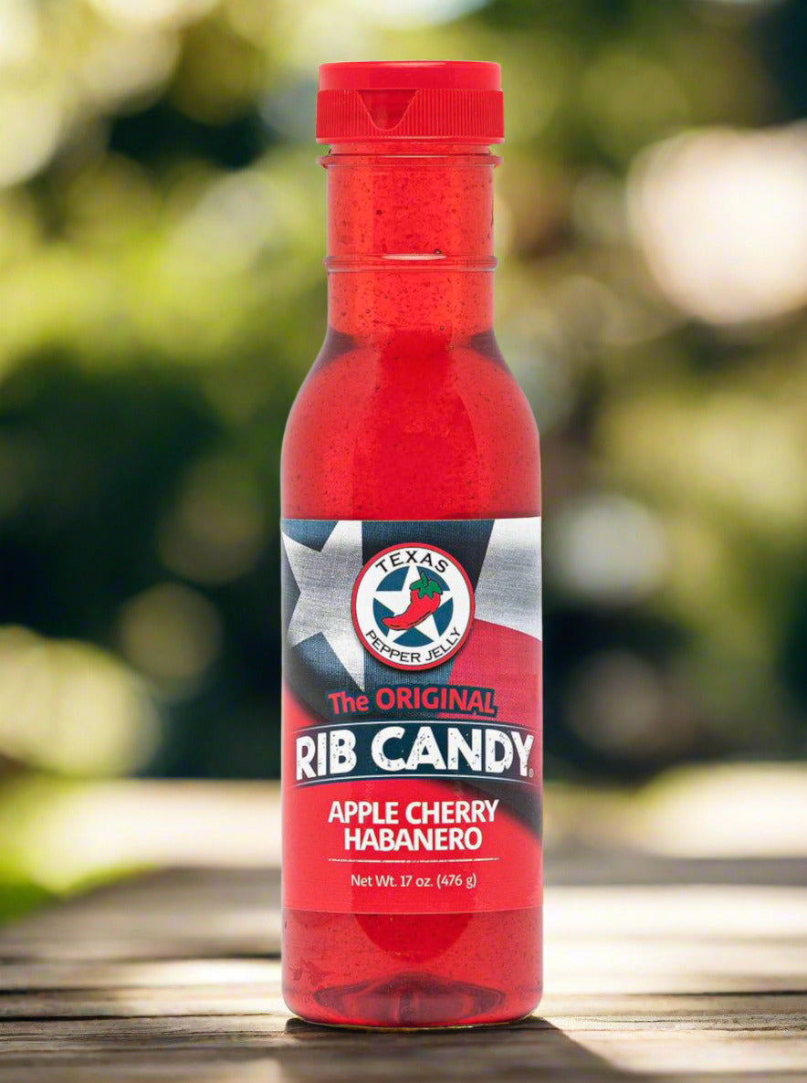 A picture of a plastic bottle of Texas Pepper Jelly's "The Original Rib Candy", Apple Cherry Habanero flavor. The bottle is cylindrical and clear & the top is covered by a plastic red flip top. The label reads: THE ORIGINAL RIB CANDY APPLE CHERRY HABANERO, NET WT 17 OZ in red and white text. Behind the text is the Texas flag in red, white, and blue. Inside the bottle is a bright red thick fluid, with air bubbles suspended in the fluid. The background shows the bottle on a wooden table in a backyard.