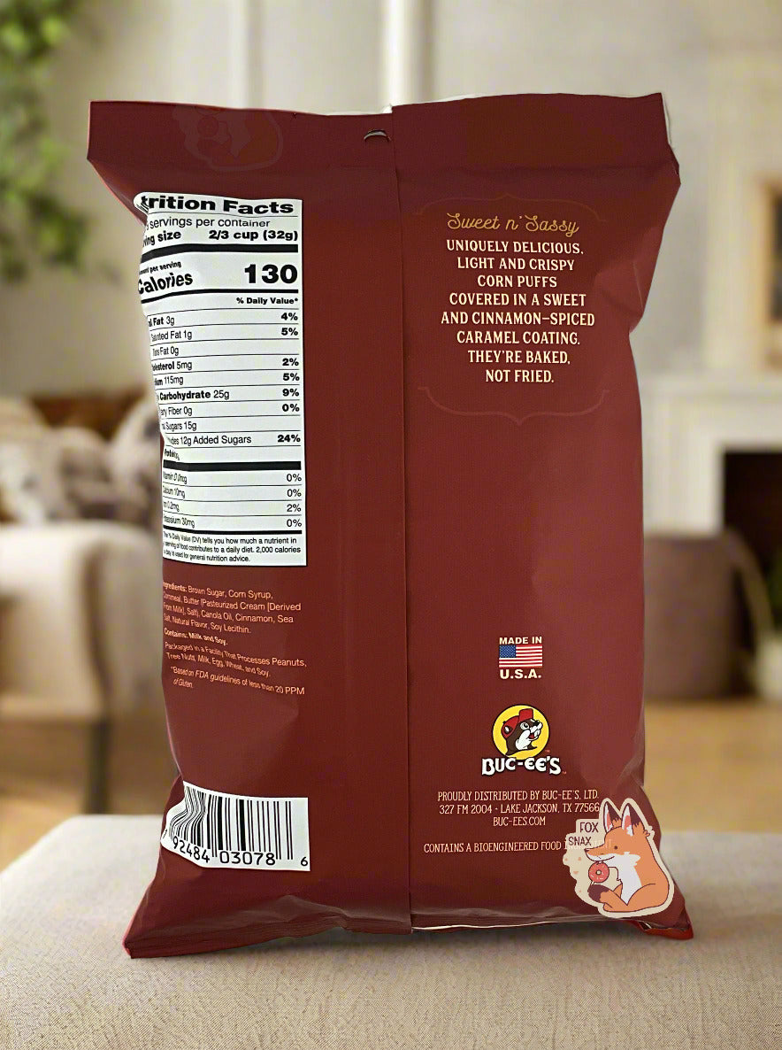 A picture of the back of a opaque, brown and white bag of Buc-ee's Cinnamon Sweet Beaver Nuggets. The bag reads: Sweet n' Sassy, Uniquely delicious, Light And Crispy, Corn Puffs covered in a sweet and cinnamon-spiced caramel coating.. They're baked not fried. The bag is sitting on a white couch in a clean living room; dark plants can be seen in the background.