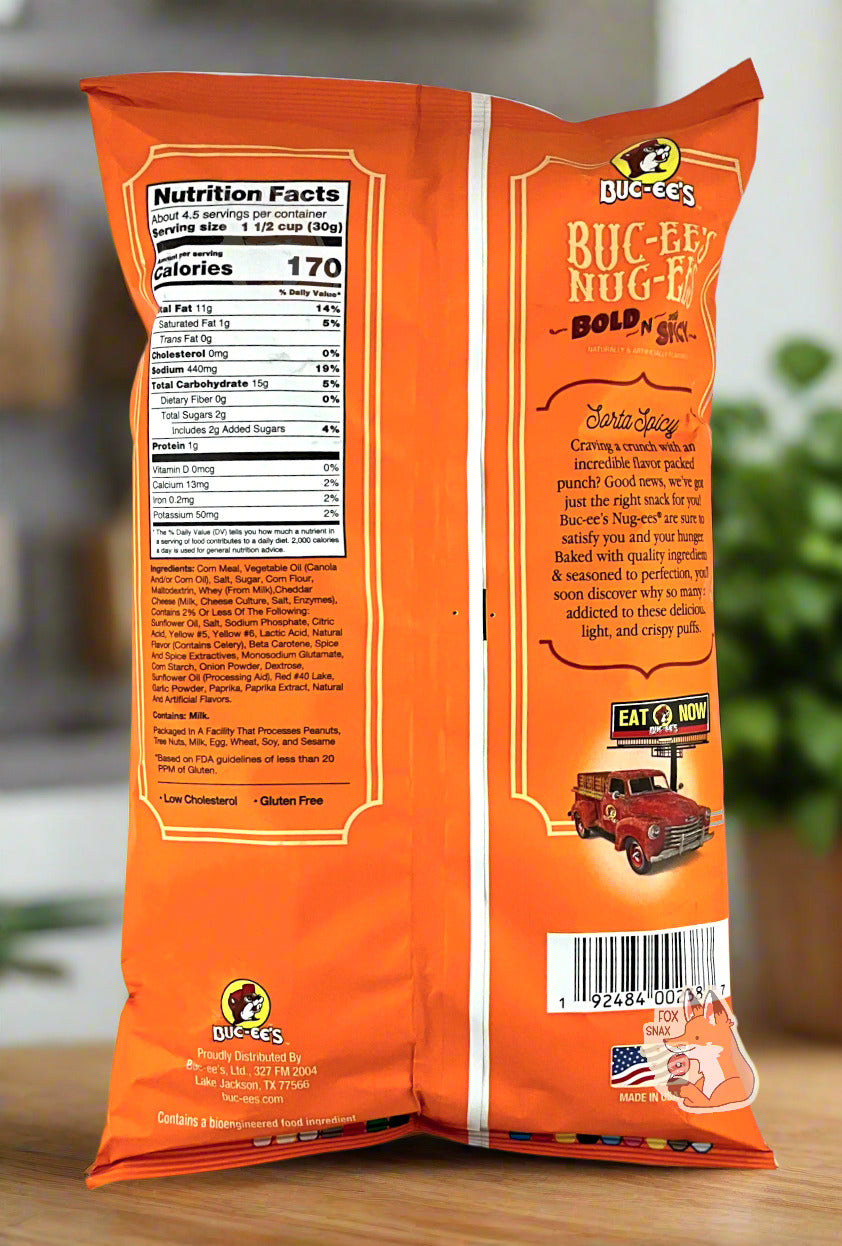 A picture of the back of a opaque, orange and yellow bag of Buc-ee's Bold N Sorta Spicy Nug-ee's. The bag reads: Sorta Spicy, Craving a crunch with incredible flavor packed punch? Good news, we've got just the right snack for you! Buc-ee's Nug-ees(r) are sure to satisfy you and your hunger. Baked with quality ingredients & seasoned to perfection, you'll soon discover why so many are addicted to these delicious, light, and crispy puffs. The background is of a nice bright kitchen with green plants.