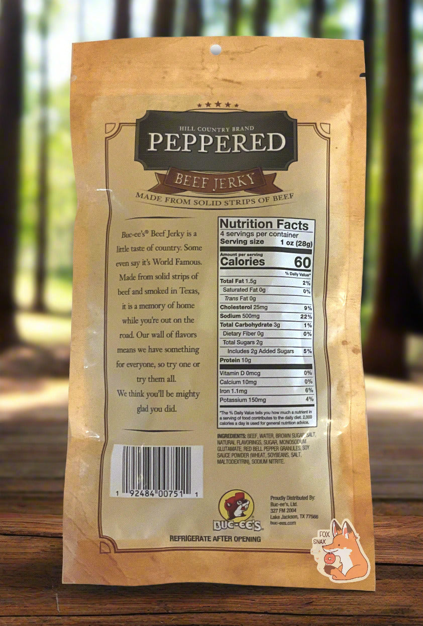 A picture of the back of a tan plastic bag of Buc-ee's Hill Country Brand Peppered Beef Jerky. The label underneath reads 'Buc-ee's Beef Jerky is a little taste of country. Some even say it's World Famous. Made from solid strips of beef and smoked in Texas, it is a memory of home while you're out on the road. Our wall of flavors means we have something for everyone, so try one or try them all. We think you'll be mighty glad you did.' The bag is sitting on a rustic picnic table in a dense forest.
