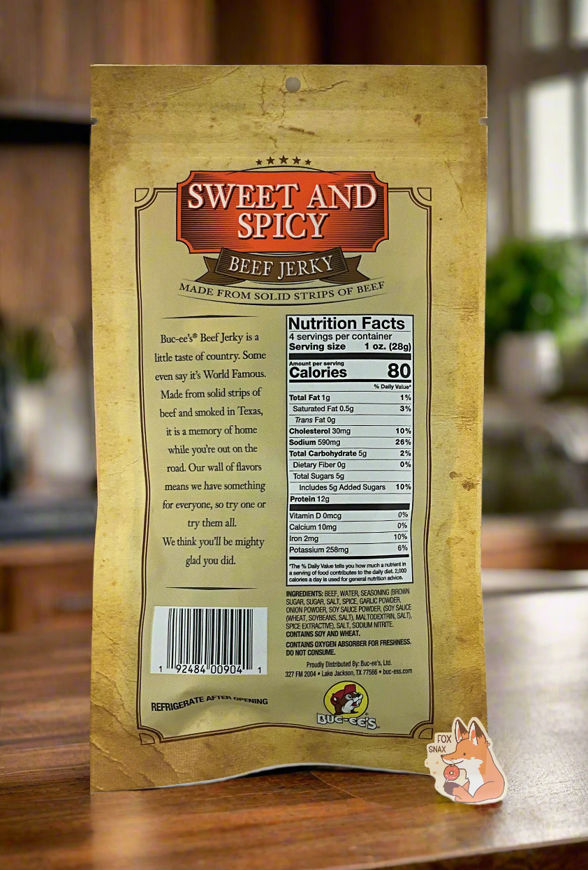 A picture of the back of a tan plastic bag of Buc-ee's Sweet And Spicy Beef Jerky. The label underneath reads 'Buc-ee's Beef Jerky is a little taste of country. Some even say it's World Famous. Made from solid strips of beef and smoked in Texas, it is a memory of home while you're out on the road. Our wall of flavors means we have something for everyone, so try one or try them all. We think you'll be mighty glad you did.' The background is of a fancy, dark-wood paneled kitchen with green plants.
