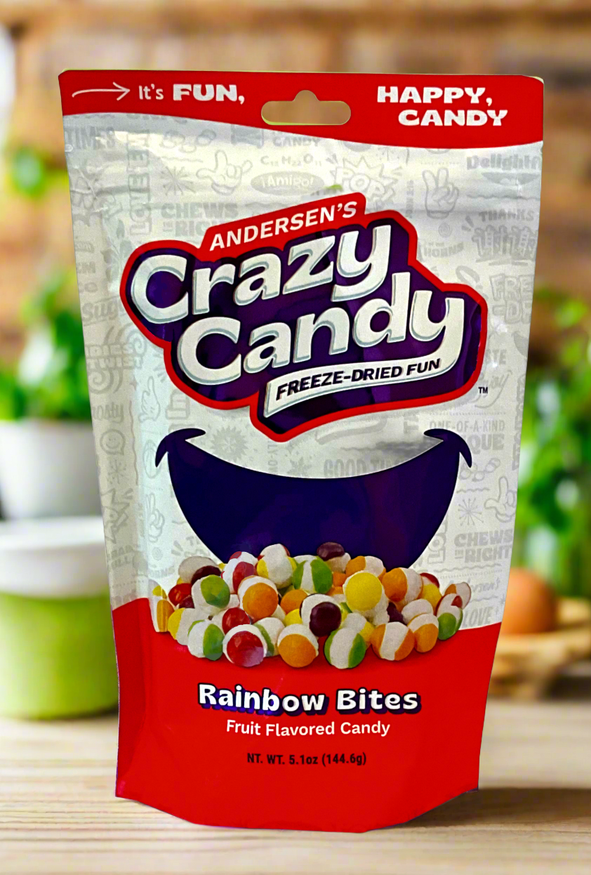 A large bag of Andersen's Crazy Candy Rainbow Bites. The front of the bag also says Freeze-Dried Fun and Fruit Flavored Candy, Net Weight 5.1 oz. It's Fun, Happy, Candy. The top and bottom of the bag are red, and the background is a large purple smile against a white background. Behind the bag is a blurred kitchen shot: the bag sits on a wooden table, and plants can be seen in the background.