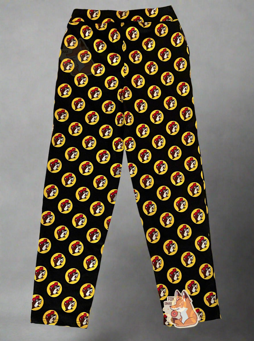 A picture of a pair of black pants. The pants are covered in Buc-ee's logos: a grinning beaver wearing a red ballcap, backlit by a yellow circle. A black drawstring can be seen at the waist of the pants. The background is a white-grey gradient.