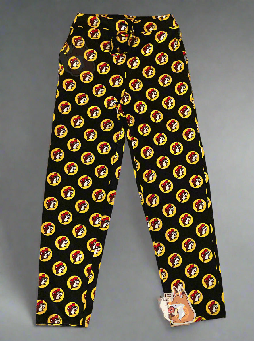 A picture of a pair of black pants. The pants are covered in Buc-ee's logos: a grinning beaver wearing a red ballcap, backlit by a yellow circle. A black drawstring can be seen at the waist of the pants. The background is a white-grey gradient.