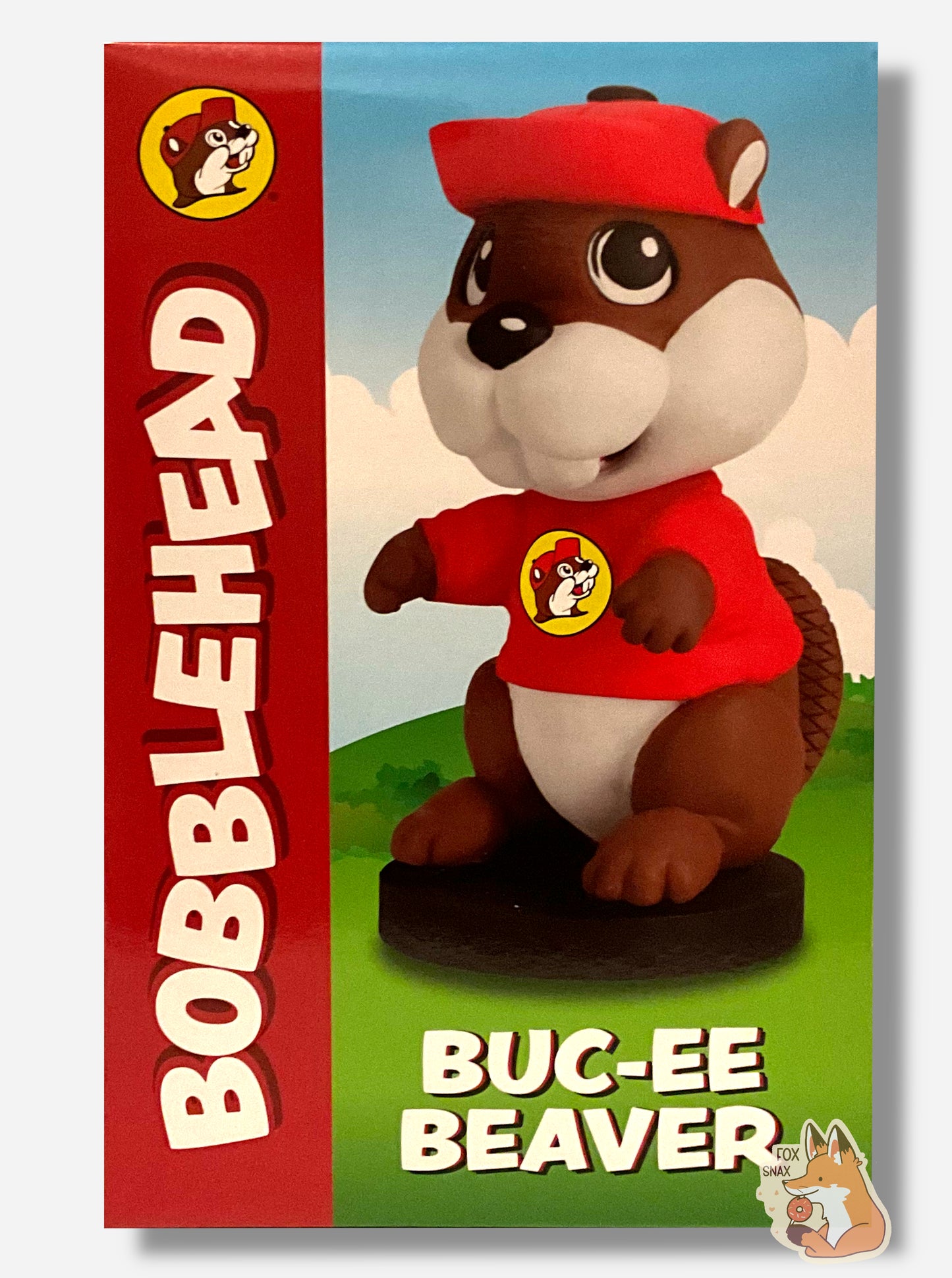 A picture of the front of a box for a Buc-ee The Beaver Bobblehead. It shows an adorable plastic beaver with a big fat beaver head; Buc-ee is wearing his usual red ball cap and Buc-ee's shirt. and has his arms in front of his body. He is standing on a small circular black pedestal; his tail can be seen behind him. The background is of a green cartoon meadow.