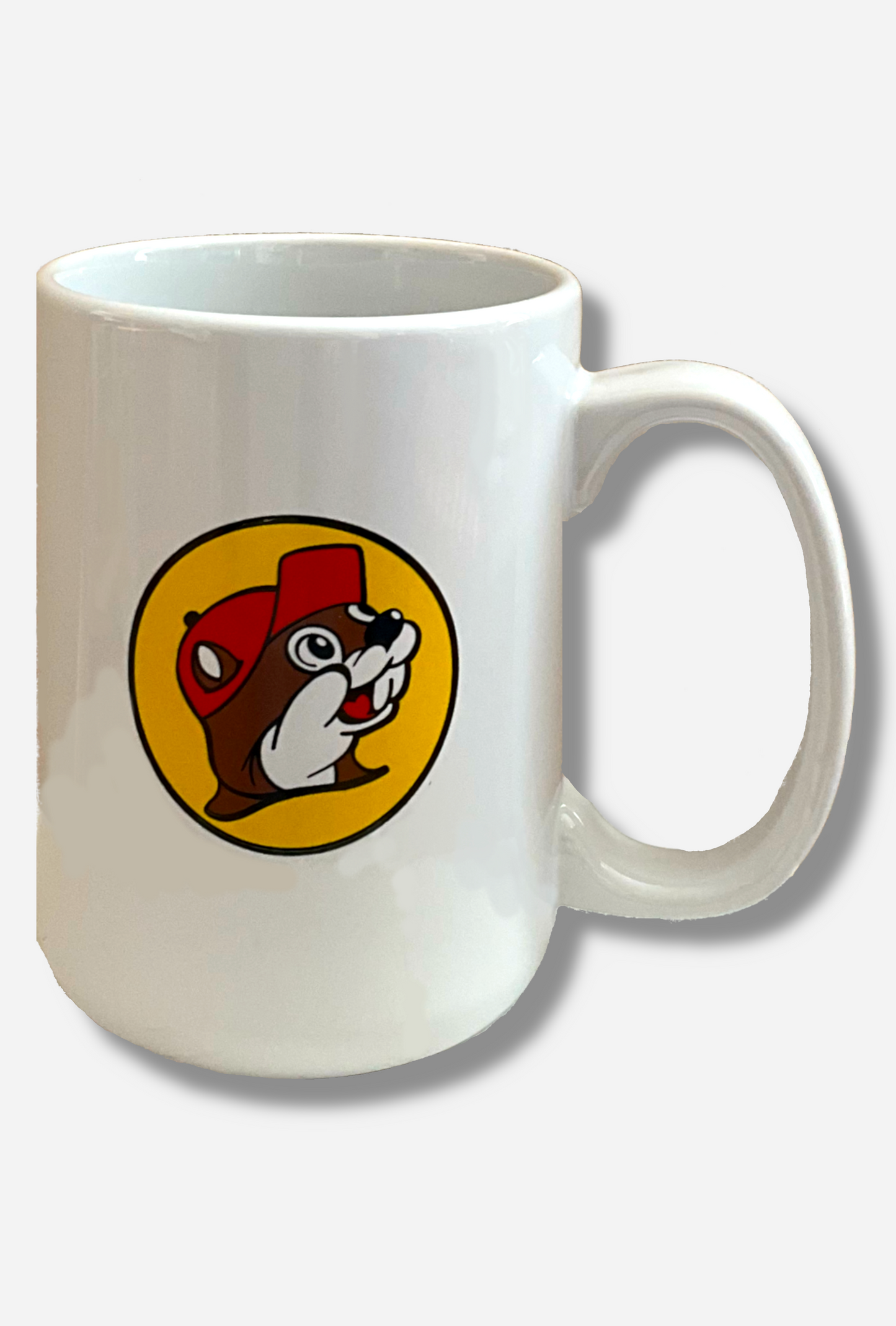 A picture of a white ceramic coffee mug, with a white handle. The middle of the mug has the Buc-ee's logo on it, which is a picture of a grinning beaver head with brown and white fur, wearing a red ballcap. The background behind the beaver is a yellow circle, outlined in black.
