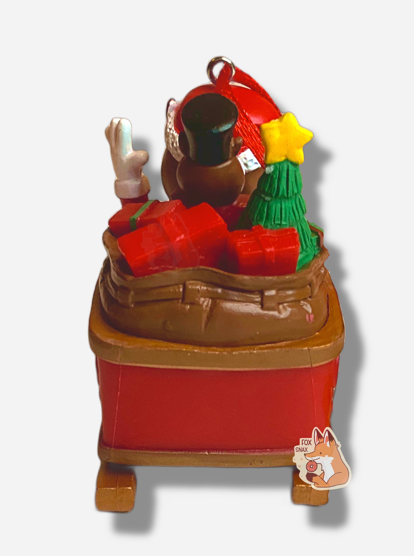 A picture of the back of a bright, plastic, Christmas ornament. The ornament is of Buc-ee the Beaver - a smiling beaver wearing a red Christmas stocking cap and Santa outfit. Buc-ee is riding in a gold and red sleigh; his left arm is raised in a greeting. A red ribbon tied to a very small hook is poking out of Buc-ee's head. In the back of the sleigh are a green Christmas tree with a yellow star, a beaver "nutcracker", and brightly colored present boxes.
