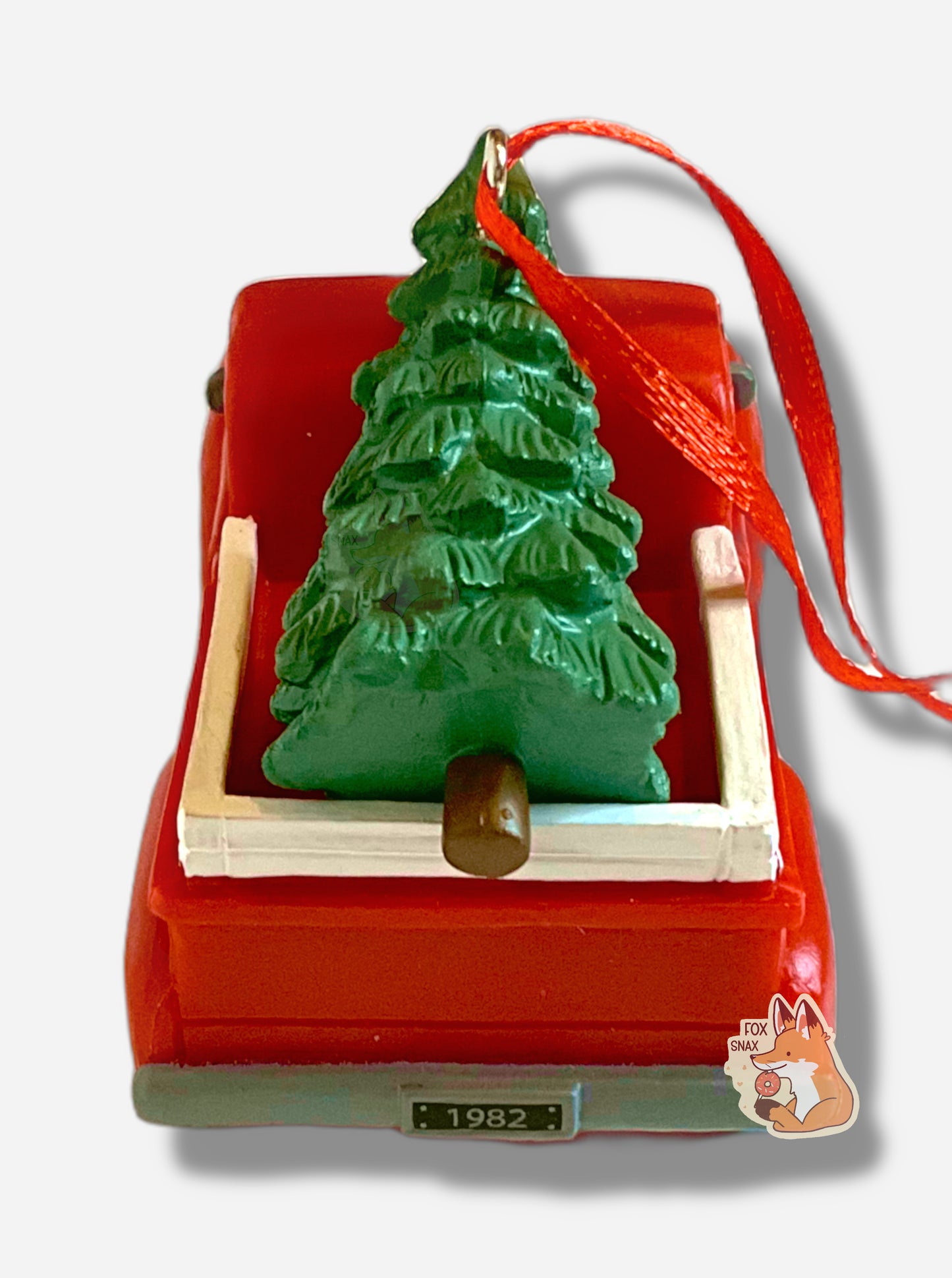 A picture of the side of a bright, plastic, Christmas ornament. The ornament is of Buc-ee's the Beaver - a smiling beaver wearing a red Christmas stocking cap. Buc-ee is driving a red pickup truck, with a green pine tree in the bed. In this picture, the back (bed) of the pickup truck can be seen, and has a grey/black license plate that reads 1982.