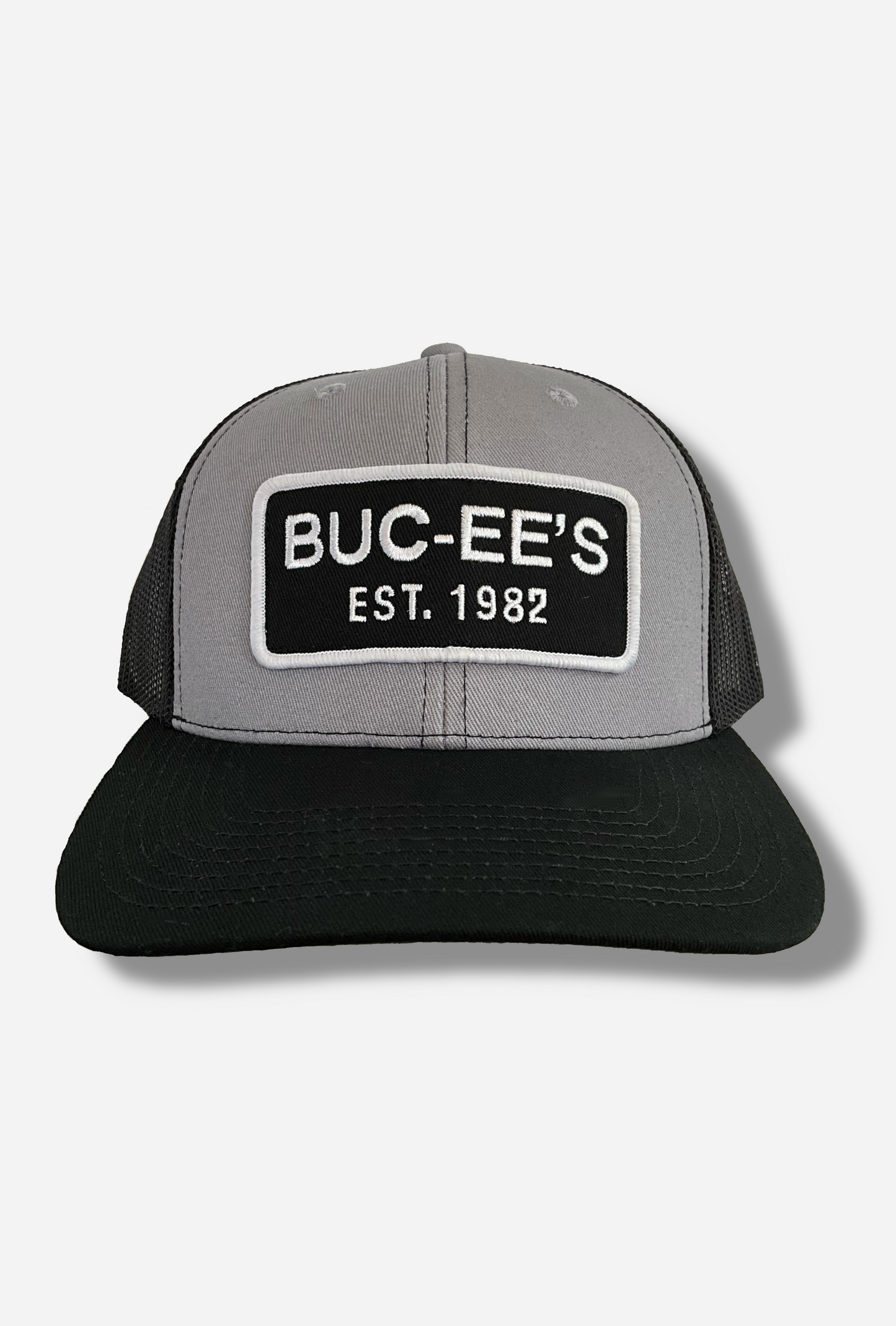 A picture of a grey and black ball cap. The front of the cap is grey, and has a large white and black rectangular patch that reads BUC-EE'S, and underneath that, EST. 1982. The brim of the cap is black. The back and sides of the cap are a black plastic mesh.