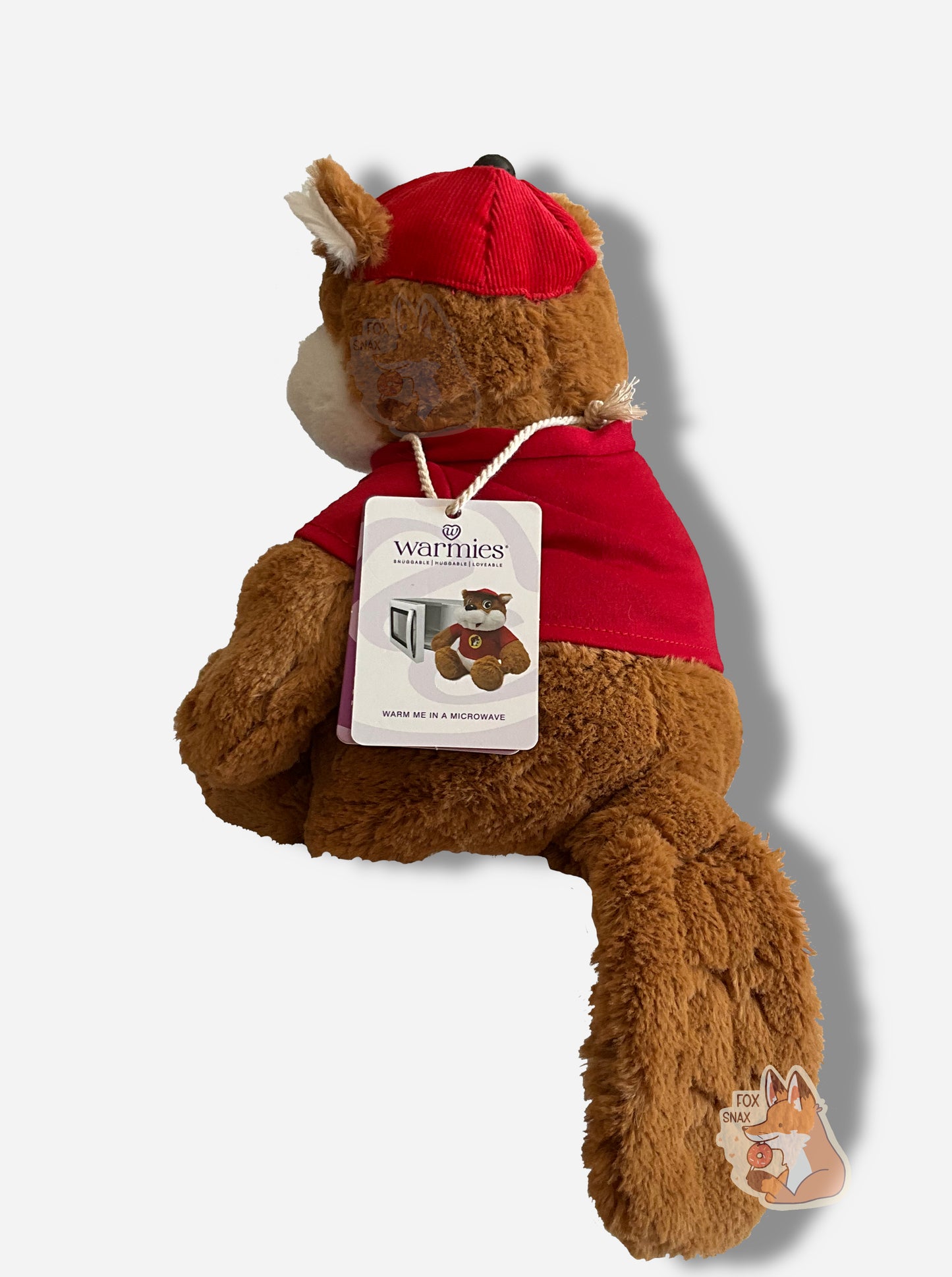 A picture of the back of an adorable Buc-ee the Beaver warmie sitting down. Buc-ee has brown and white fur, and is wearing a red cap. His shirt is a red shirt, with a Buc-ee's logo on it. Around his neck is a white cord with a paper tag on it that reads "WARMIES, SNUGGABLE | HUGGABLE | LOVABLE", then "WARM ME IN A MICROWAVE". The tag has a picture of the Buc-ee warmie sitting next to an open white microwave.