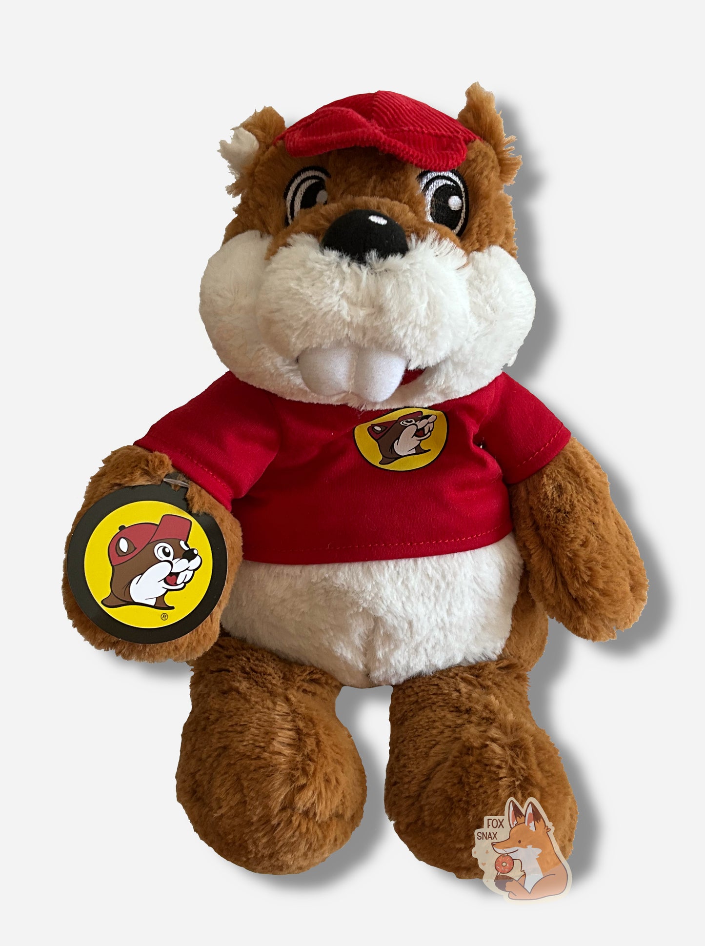 A picture of an adorable Buc-ee the Beaver warmie sitting. Buc-ee has brown and white fur, and is wearing a red cap. His shirt is a red shirt, with a Buc-ee's logo on it.