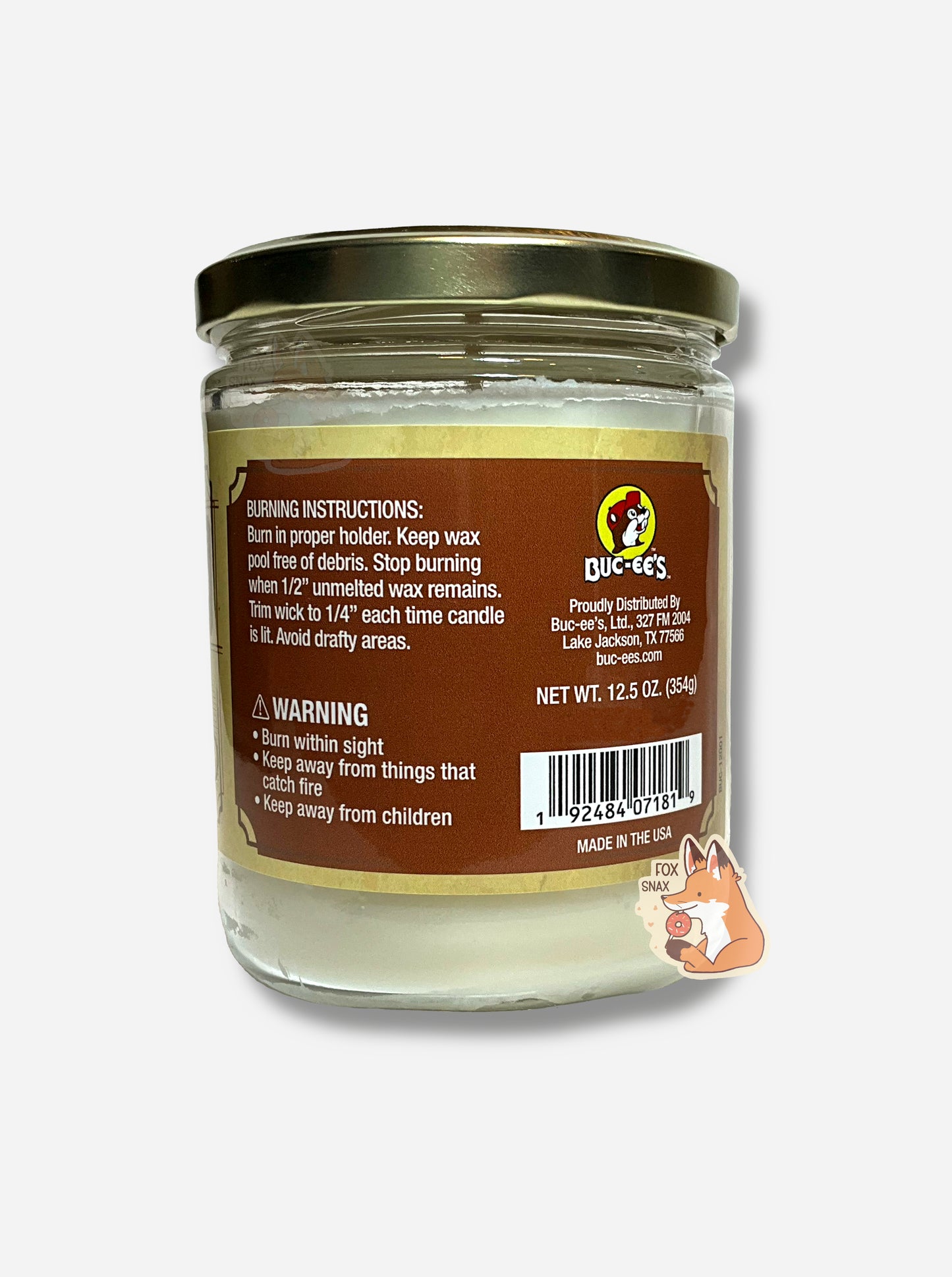 A picture of a white/yellow candle in a cylindrical glass container. The lid of the container is a gold colored metal lid. The text on a brown sticker on the back reads BURNING INSTRUCTIONS: Burn in proper holder. Keep wax pool free of debris. Stop burning when 1/2" unmelted wax remains. Trim wick to 1/4" each time candle is lit. Avoid drafty areas. Net Wt 12.5 Oz. MADE IN THE USA