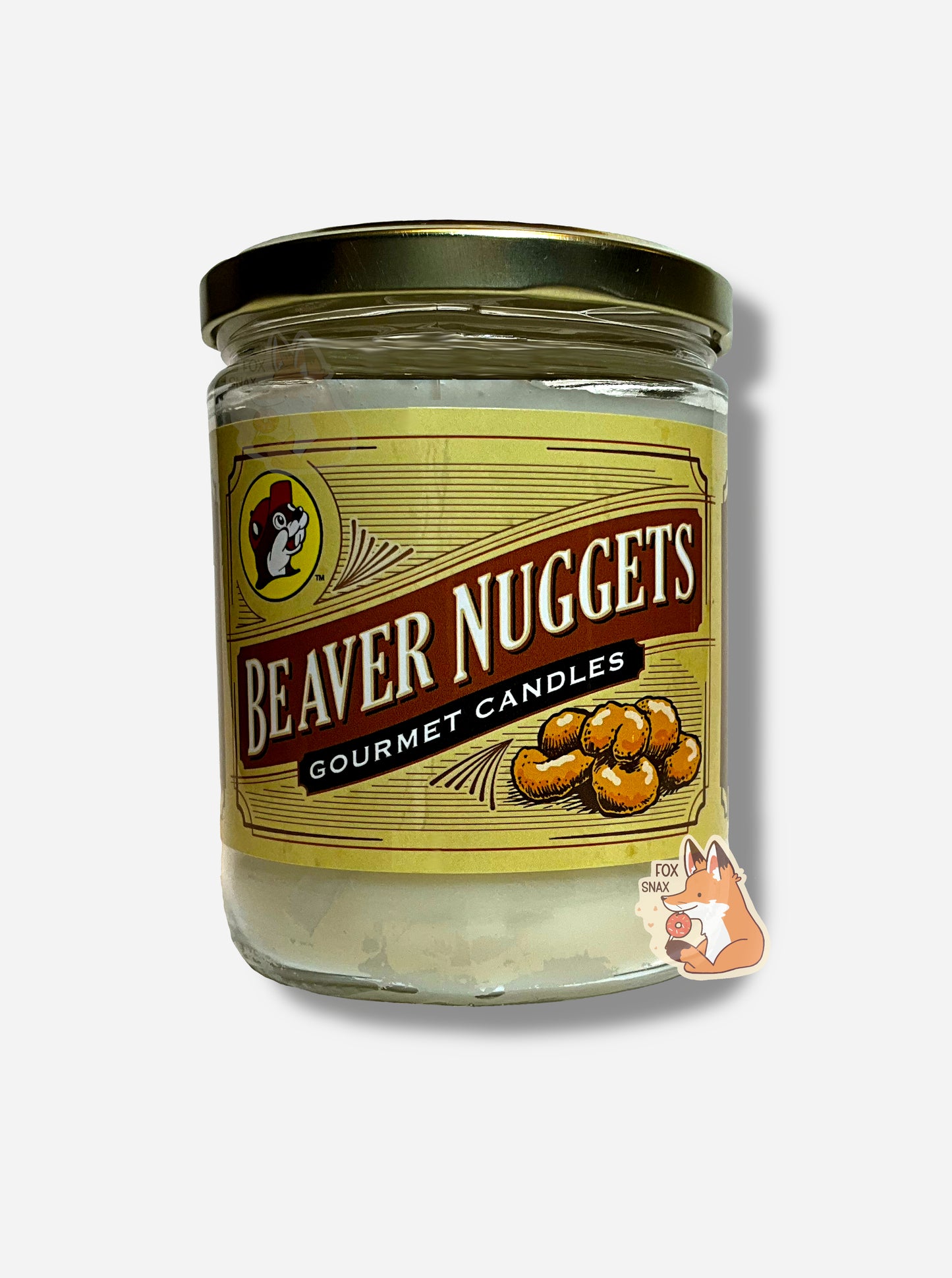 A picture of a white/yellow candle in a cylindrical glass container. The lid of the container is a gold colored metal lid. The front of the candle has a yellow sticker with the Buc-ee's logo, then underneath that, BEAVER NUGGETS, then GOURMET CANDLES, then a picture of six beaver nuggets.