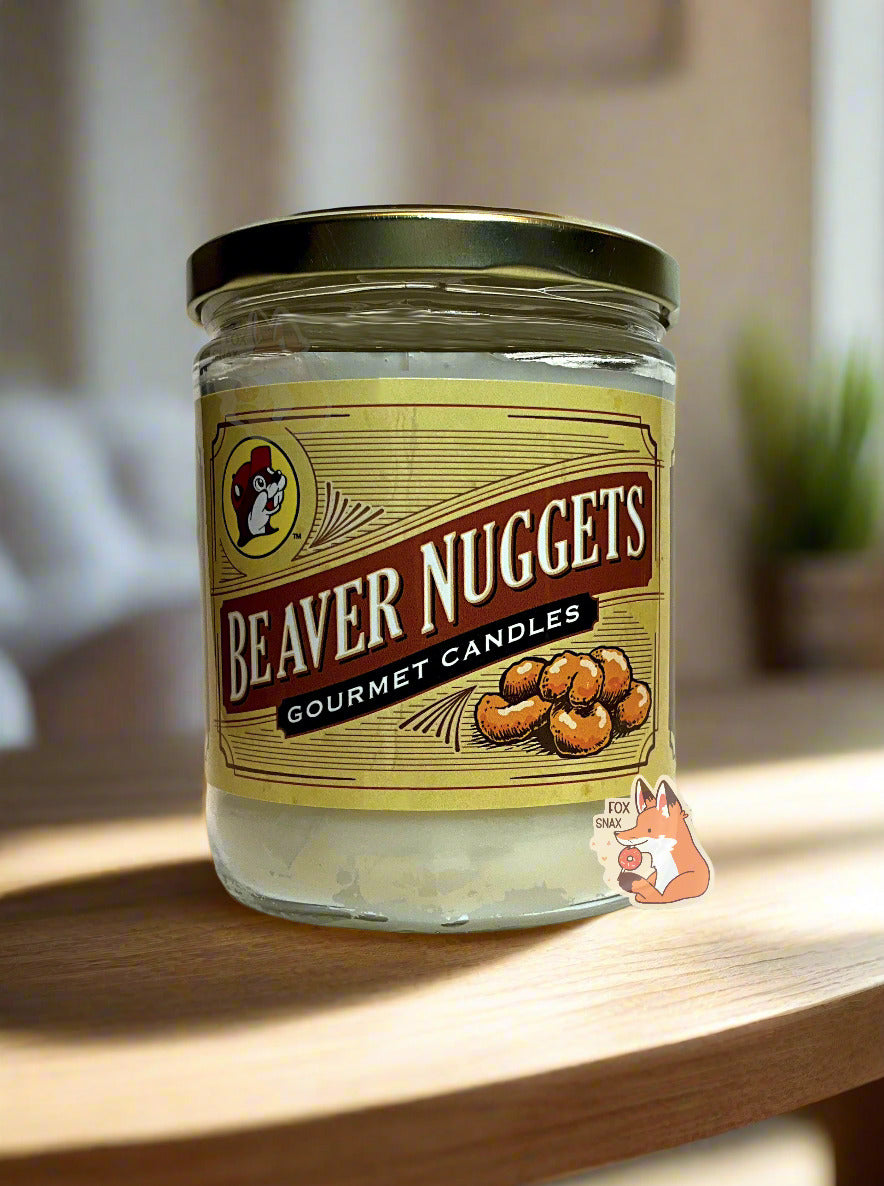 A picture of a white/yellow candle in a cylindrical glass container. The lid of the container is a gold colored metal lid. The front of the candle has a yellow sticker with the Buc-ee's logo, then underneath that, BEAVER NUGGETS, then GOURMET CANDLES, then a picture of six beaver nuggets. The candle is sitting on a wooden table; sunlight overlaps the candle and a small plant can be seen in the background.