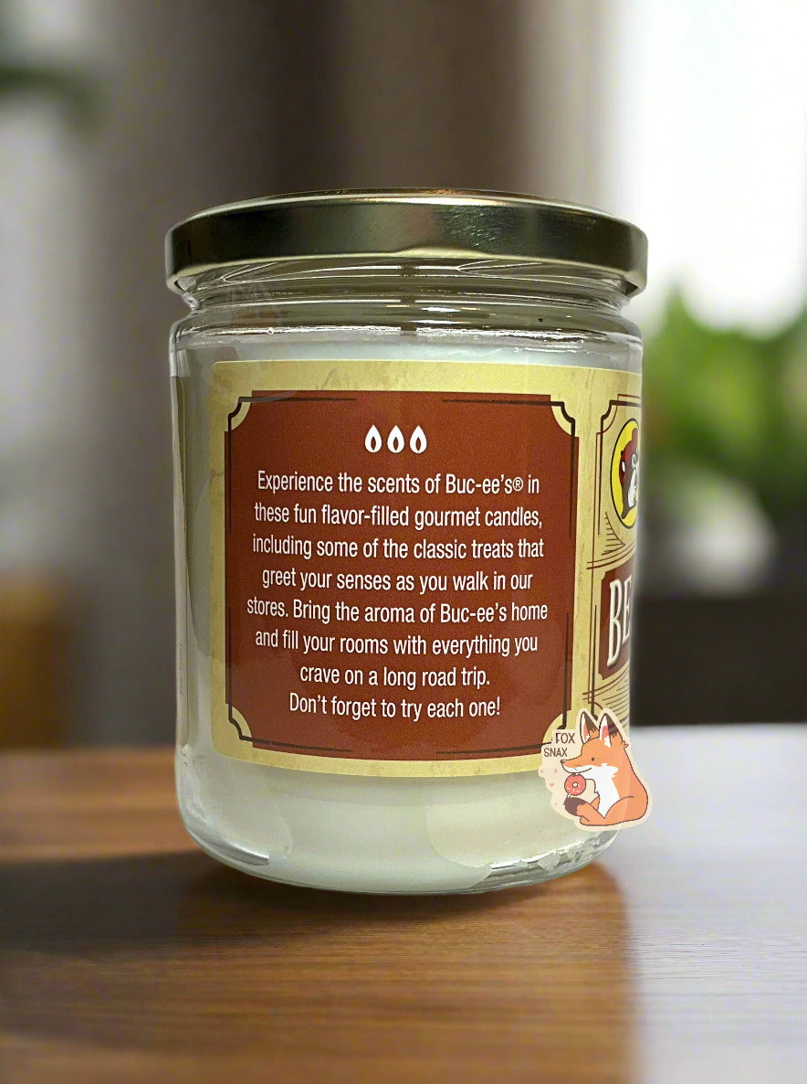 A picture of a white/yellow candle in a cylindrical glass container. The lid of the container is a gold colored metal lid. The text on a brown sticker on the side reads: Experience the scents of Buc-ee's in these fun flavor-filled gourmet candles, including some of the classic treats that greet your senses as you walk in our stores. Bring the aroma of Buc-ee's home and fill your rooms with everything you crave on a long road trip. Don't forget to try each one! The candle is sitting on a wooden table.