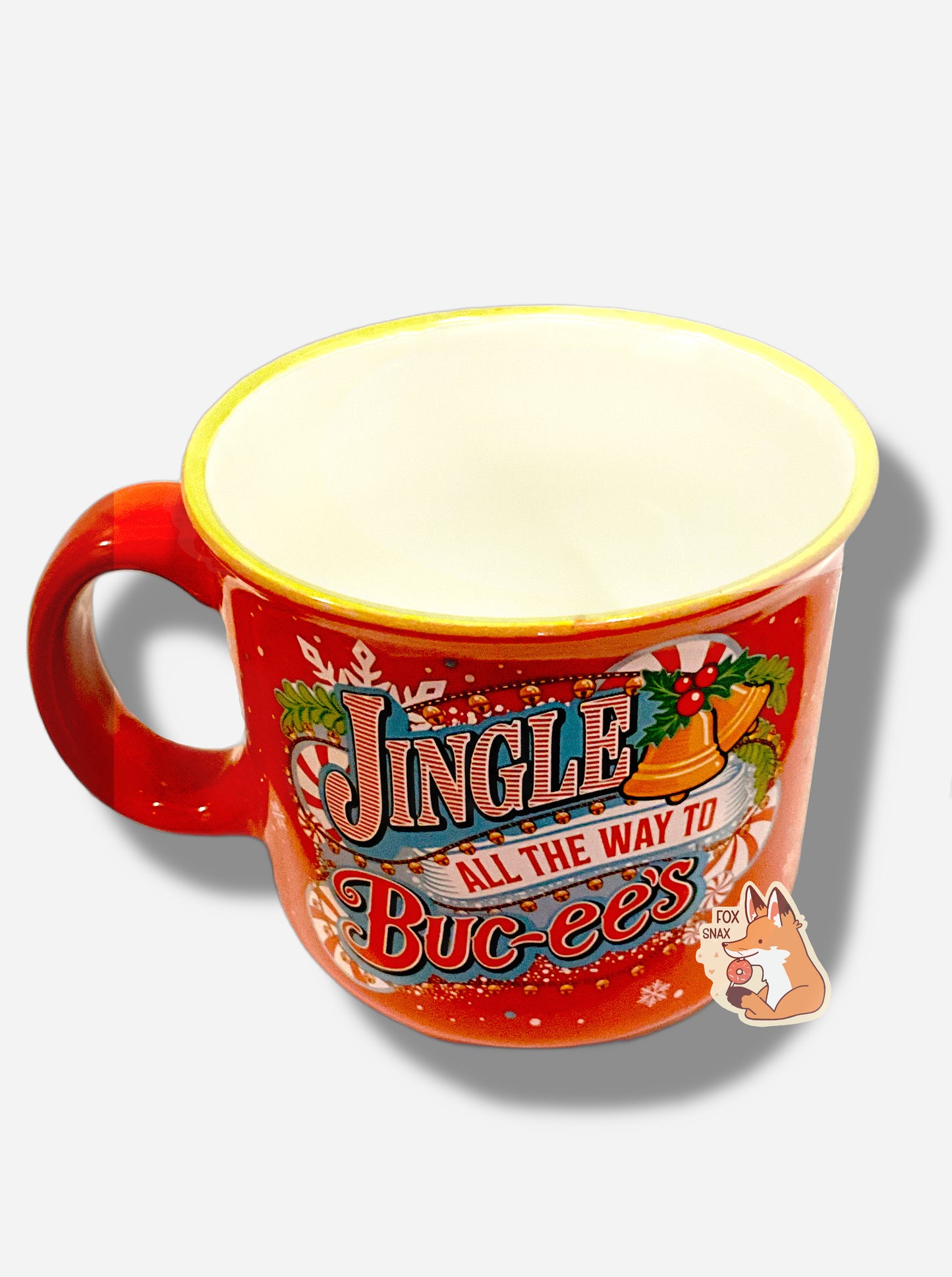 A festive, red ceramic mug, covered in white snowflakes and red/white peppermints. The mug is decorated with the words JINGLE ALL THE WAYS TO BUC-EE'S. These words are framed with yellow jingle bells, holly leaves and berries, peppermints, and snowflakes. The handle of the mug is red. The inside of the mug is white.