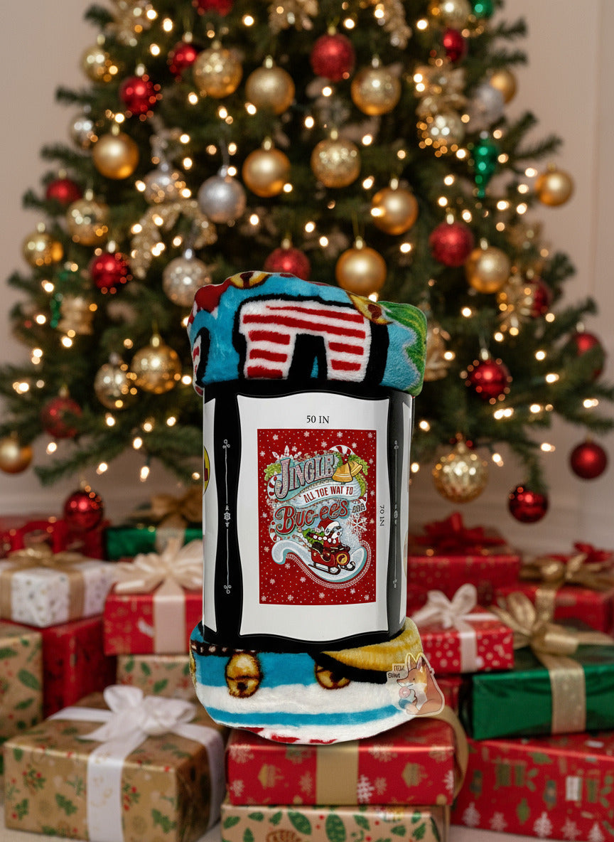 A picture of the side of a soft white, blue, blank, yellow, green, and red (!) blanket featuring Buc-ee the Beaver in a red Christmas sleigh, with a green Christmas tree in the back. Buc-ee is holding the front of the sleigh, and is wearing a Christmas cap and red and white striped scarf. The background is in red, framed by white snowflakes at the top, and reads JINGLE ALL THE WAY TO BUC-EE'S. The blanket is sitting on a pile of presents underneath a beautiful Christmas tree.