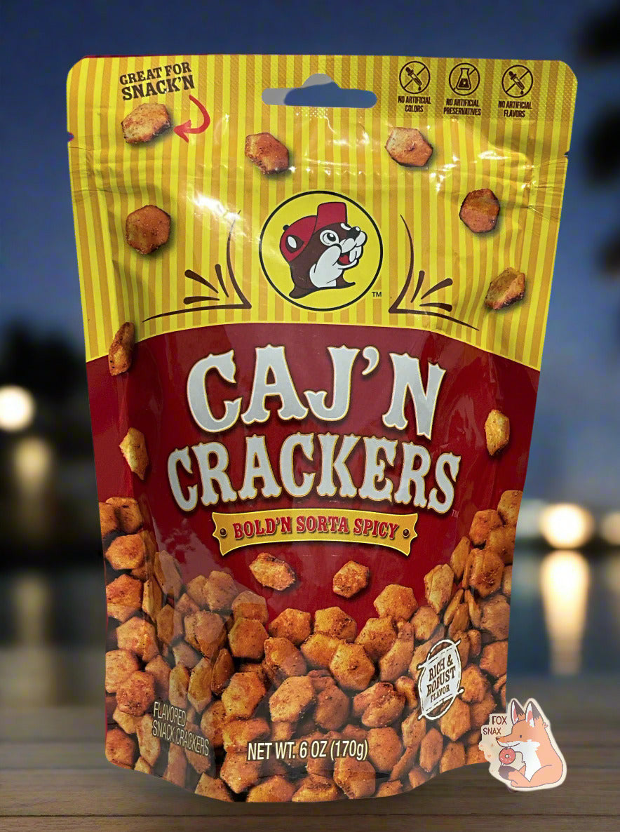 A picture of a metallic bag of Buc-ee's Caj'N Crackers and Bold'N Sorta Spicy. The logo is a dark red background on white text; a large number of reddish, savory-looking hexagonal oyster crackers are shown beneath it. At the top is a picture of the logo of Buc-ee The Beaver, and it reads GREAT FOR SNACK'N, NO ARTIFICIAL COLORS, NO ARTIFICIAL PRESERVATIVES, NO ARTIFICIAL FLAVORS and RICH AND ROBUST FLAVOR. The background is of a well-lit dock by the bayou.