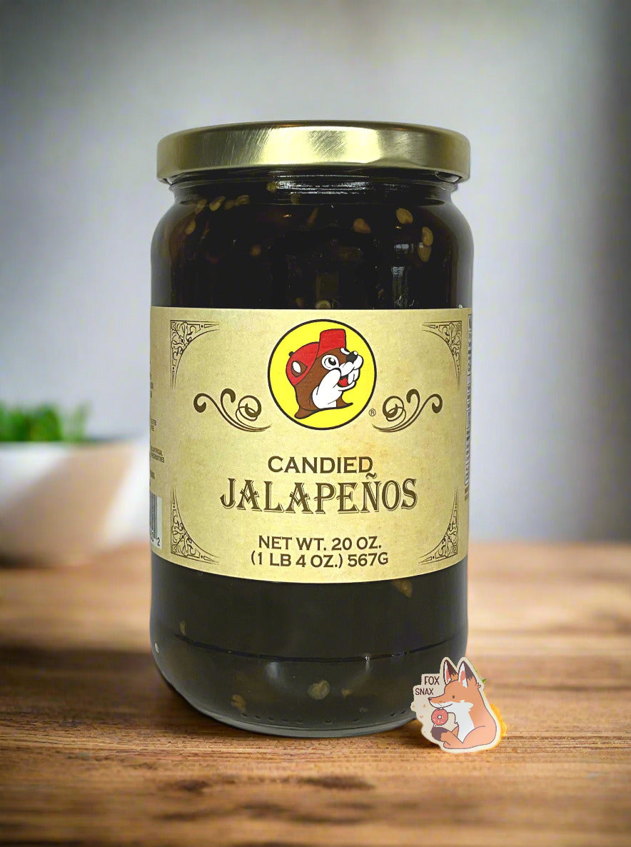 A picture of the front of a glass jar of Buc-ee's Candied Jalapeños - the label is a "old-timey" sort of white/yellow with flared edges. The middle has a giant Buc-ee the Beaver logo on it. In the jar, dark green jalapeños can be seen. The background shows the jar on the wooden table in a pretty kitchen; a white bowl and plants can be seen against a grey wall.