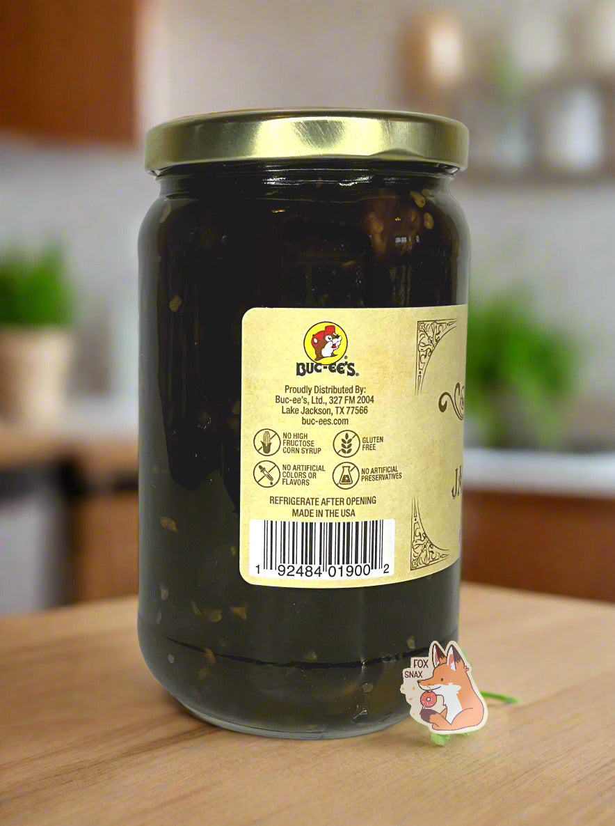 A picture of the left side of a glass jar of Buc-ee's Candied Jalapeños - the label is a "old-timey" sort of white/yellow with flared edges. The middle reads No High Fructose Corn Syrup, No Artificial Colors Or Flavors, Gluten Free, No Artificial Preservatives. In the jar, dark green jalapeños can be seen. The background shows the jar on the wooden table in a pretty kitchen; a white bowl and plants can be seen against a grey wall.