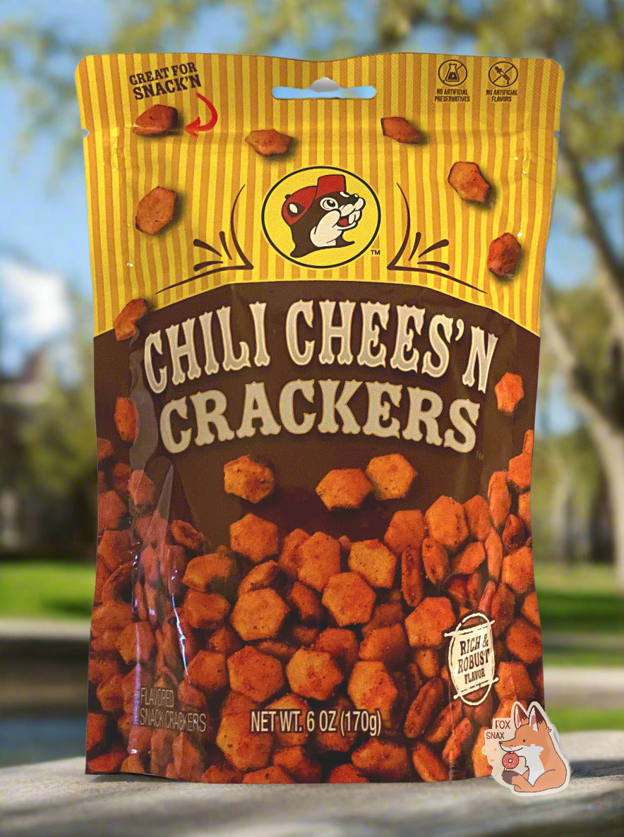 A picture of a metallic bag of Buc-ee's Chili Chees'N Crackers. The logo is a brown-red background on white text; a large number of reddish, savory-looking hexagonal oyster crackers are shown beneath it. At the top is a picture of the logo of Buc-ee The Beaver, and it reads GREAT FOR SNACK'N, NO ARTIFICIAL PRESERVATIVES, FLAVORS and RICH AND ROBUST FLAVOR. The background is of a park on a sunny spring day.