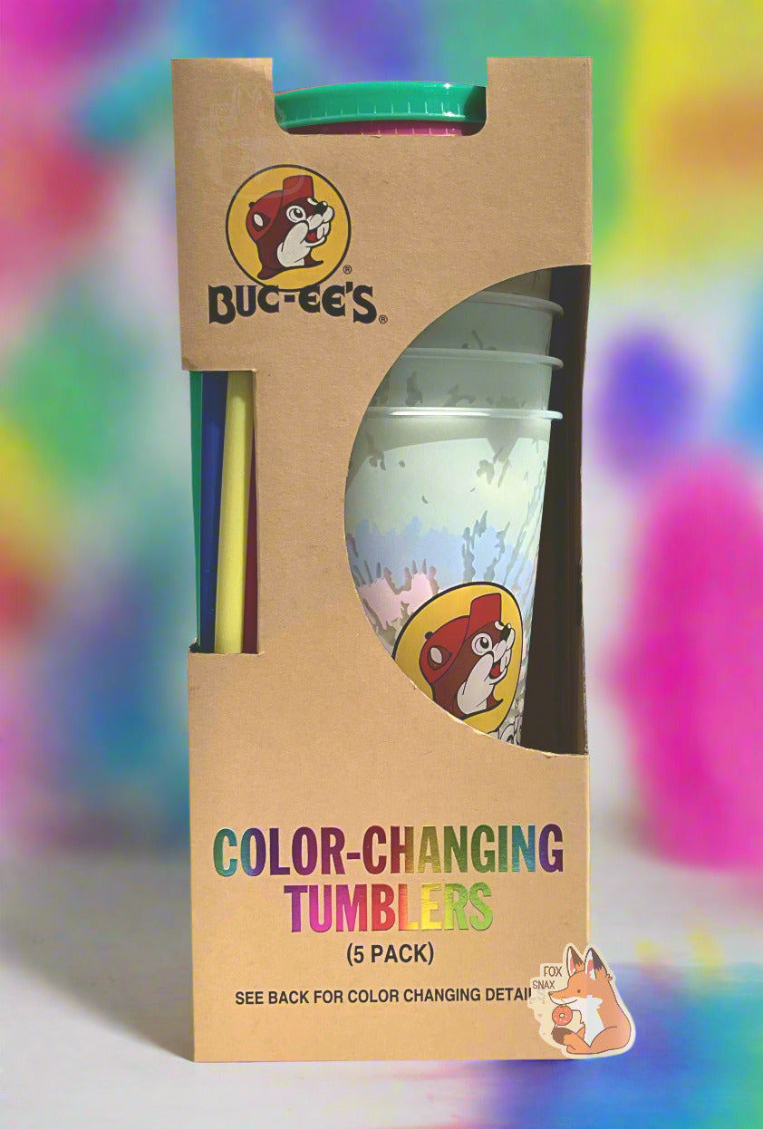 A picture of a tall, thin cardbox box. The bottom of the box reads COLOR-CHANGING TUMBLERS. The top of the box has the Buc-ee's logo. In the middle is a semicircle cut out, which exposes the plastic tumblers. Several tumblers can be seen, and they show a faded, white-ish plastic tumbler with a faint tie-dye pattern through the semicircle. Each of the tumblers is tall, plastic, and has the Buc-ee's logo on it. The background shows a groovy multicolored set of pastel hues.
