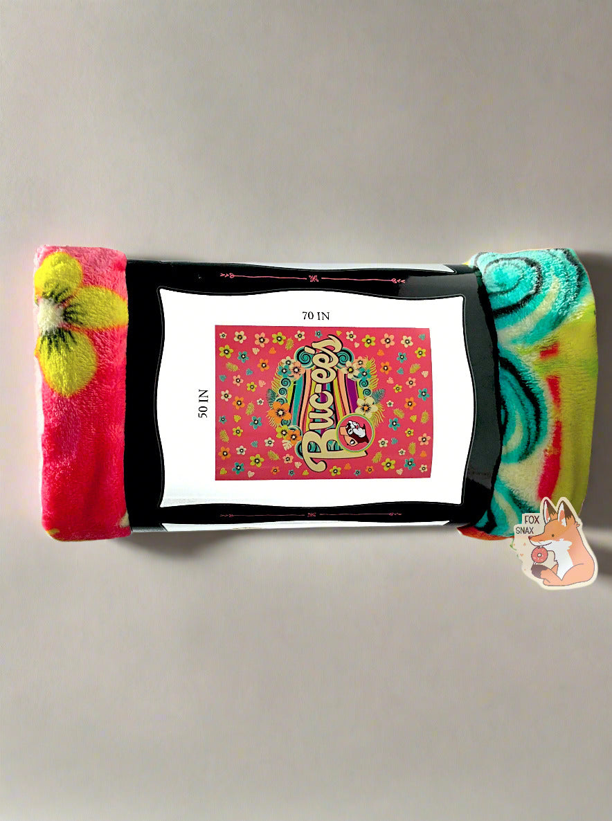 A picture of the other side of a rolled up blanket. The blanket is a multicolored (pink, yellow, orange, black, green/blue) blanket with pastel florals and a wave of soothing colors through the center. The iconic beaver makes an appearance, rocking a pink cap, with curvy white Buc-ee's text to match. The paper label surrounding the blanket reads BUC-EE'S BLANKET *SOFT AND COMFY* 50 IN x 70 IN. The background is a flat tan one, with soft sun shadows framing the wrapped blanket.