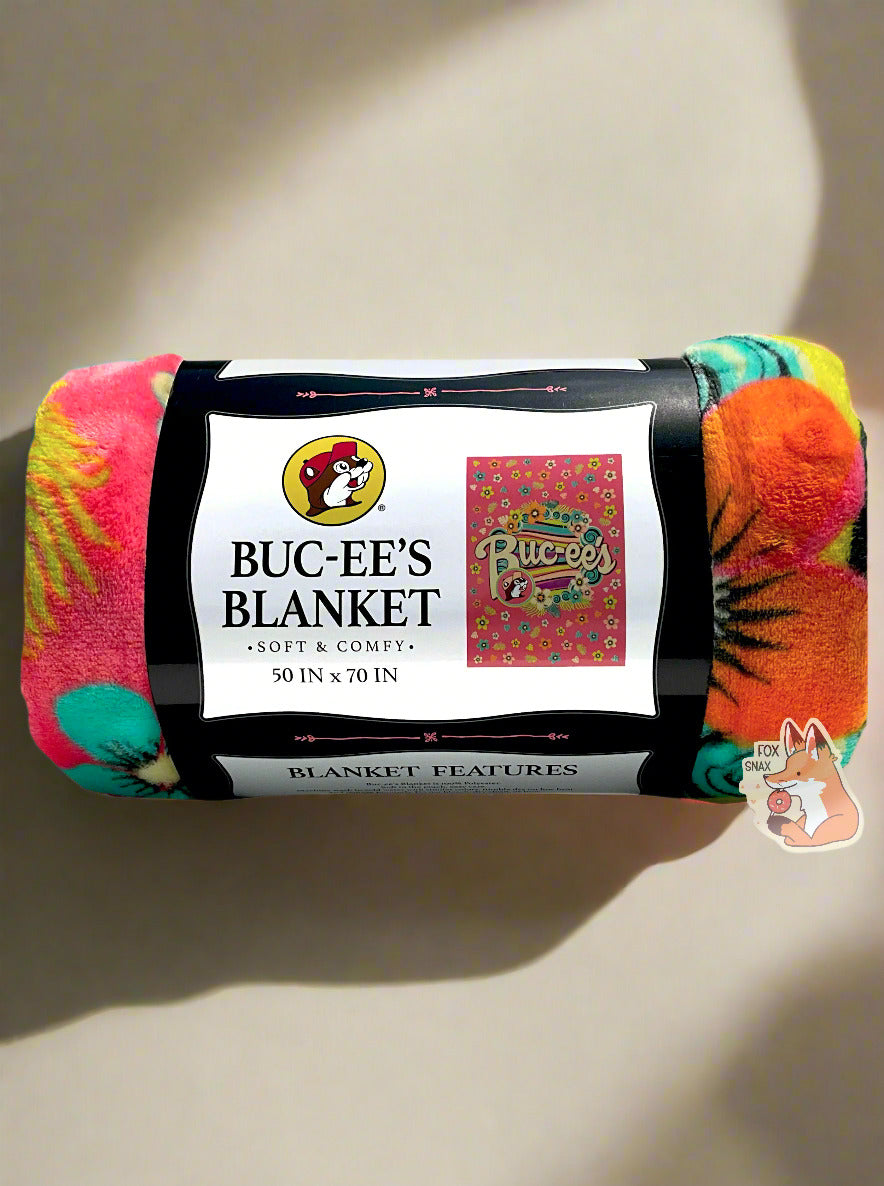 A picture of a rolled up blanket. The blanket is a multicolored (pink, yellow, orange, black, green/blue) blanket with pastel florals and a wave of soothing colors through the center. The iconic beaver makes an appearance, rocking a pink cap, with curvy white Buc-ee's text to match. The paper label surrounding the blanket reads BUC-EE'S BLANKET *SOFT AND COMFY* 50 IN x 70 IN. The background is a flat tan one, with soft sun shadows framing the wrapped blanket.