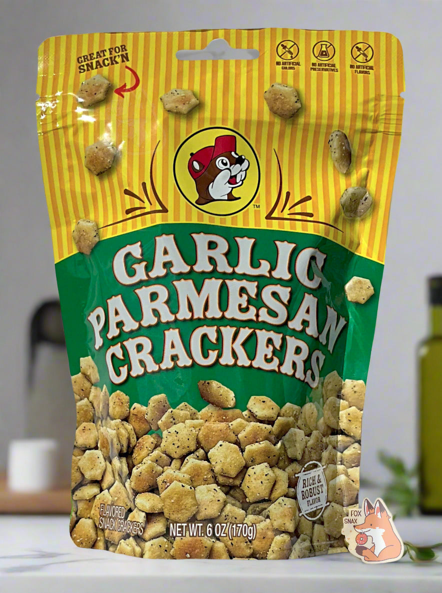 A picture of a metallic bag of Buc-ee's Garlic Parmesan Crackers. The logo is a green background on white text; a large number of savory-looking hexagonal oyster crackers are shown beneath it. At the top is a picture of the logo of Buc-ee The Beaver, and it reads GREAT FOR SNACK'N, NO ARTIFICIAL COLORS, NO ARTIFICIAL PRESERVATIVES, NO ARTIFICIAL FLAVORS and RICH AND ROBUST FLAVOR. The background shows a kitchen with grey walls; olive oil and salt containers can be seen in the background.
