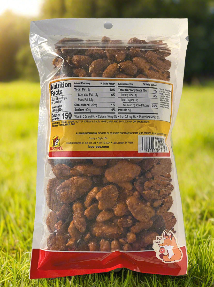 A picture of the back of a clear plastic bag of Buc-ee's Butter Toffee Pecans. Inside the bag are a large number of dried pecans, covered in brownish sugary praline material. The background behind the bag is a grassy meadow, framed by happy sunlight.