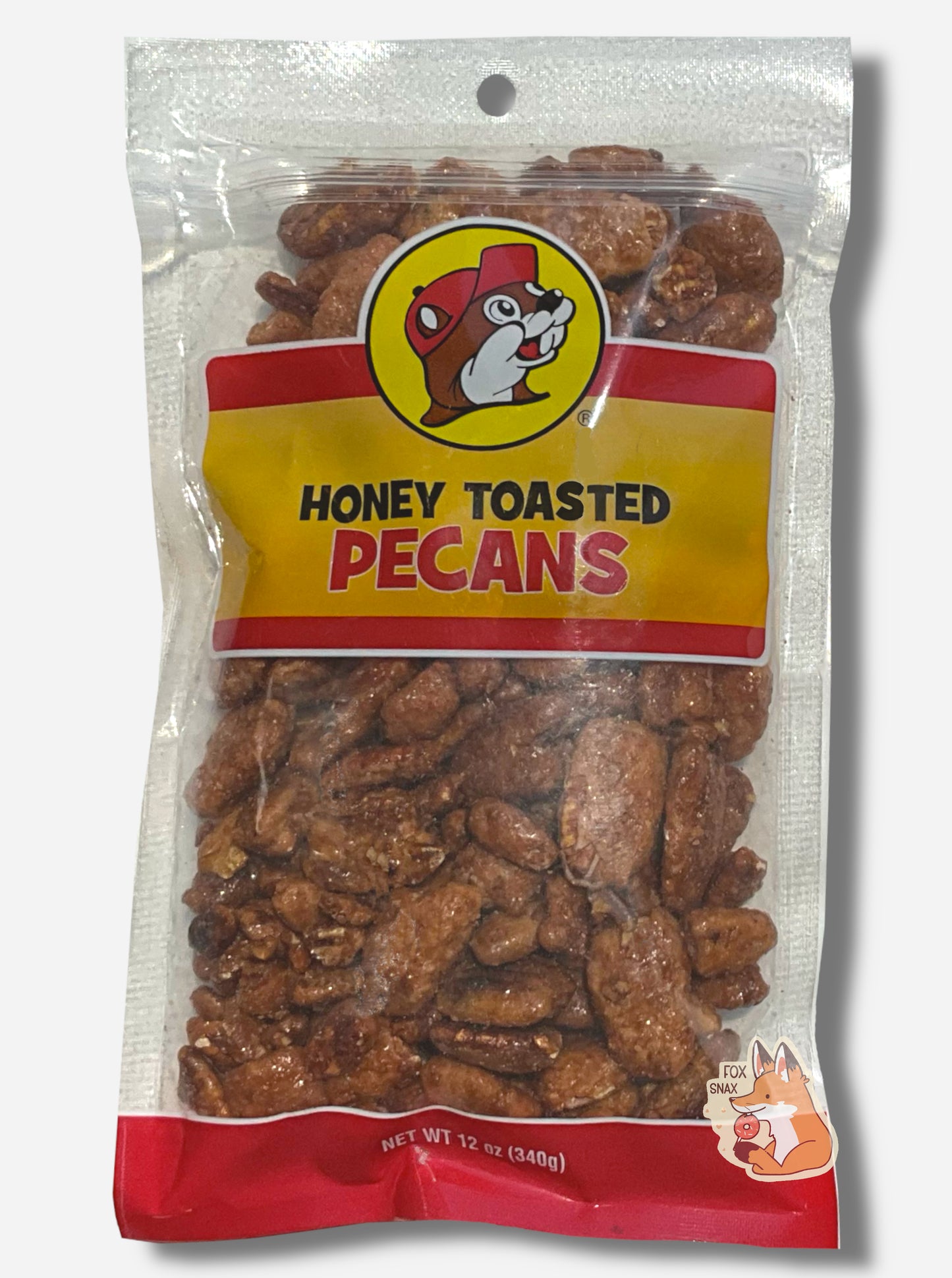 A picture of a clear plastic bag of Buc-ee's Honey Toasted Pecans. The Buc-ee's logo can be seen in the top center of the bag, then underneath that in black and red text, HONEY TOASTED PECANS. The bottom of the bag is red and reads Net Wt 12 oz. Inside the bag are a large number of toasted pecans, covered in brownish sugary material.
