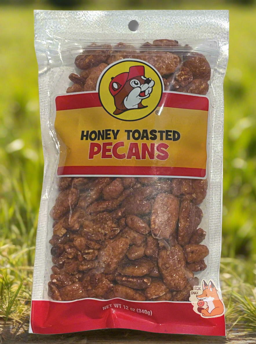 A picture of a clear plastic bag of Buc-ee's Honey Toasted Pecans. The Buc-ee's logo can be seen in the top center of the bag, then underneath that in black and red text, HONEY TOASTED PECANS. The bottom of the bag is red and reads Net Wt 12 oz. Inside the bag are a large number of dried pecans, covered in brownish sugary material. The background behind the bag is a grassy meadow, framed by happy sunlight.