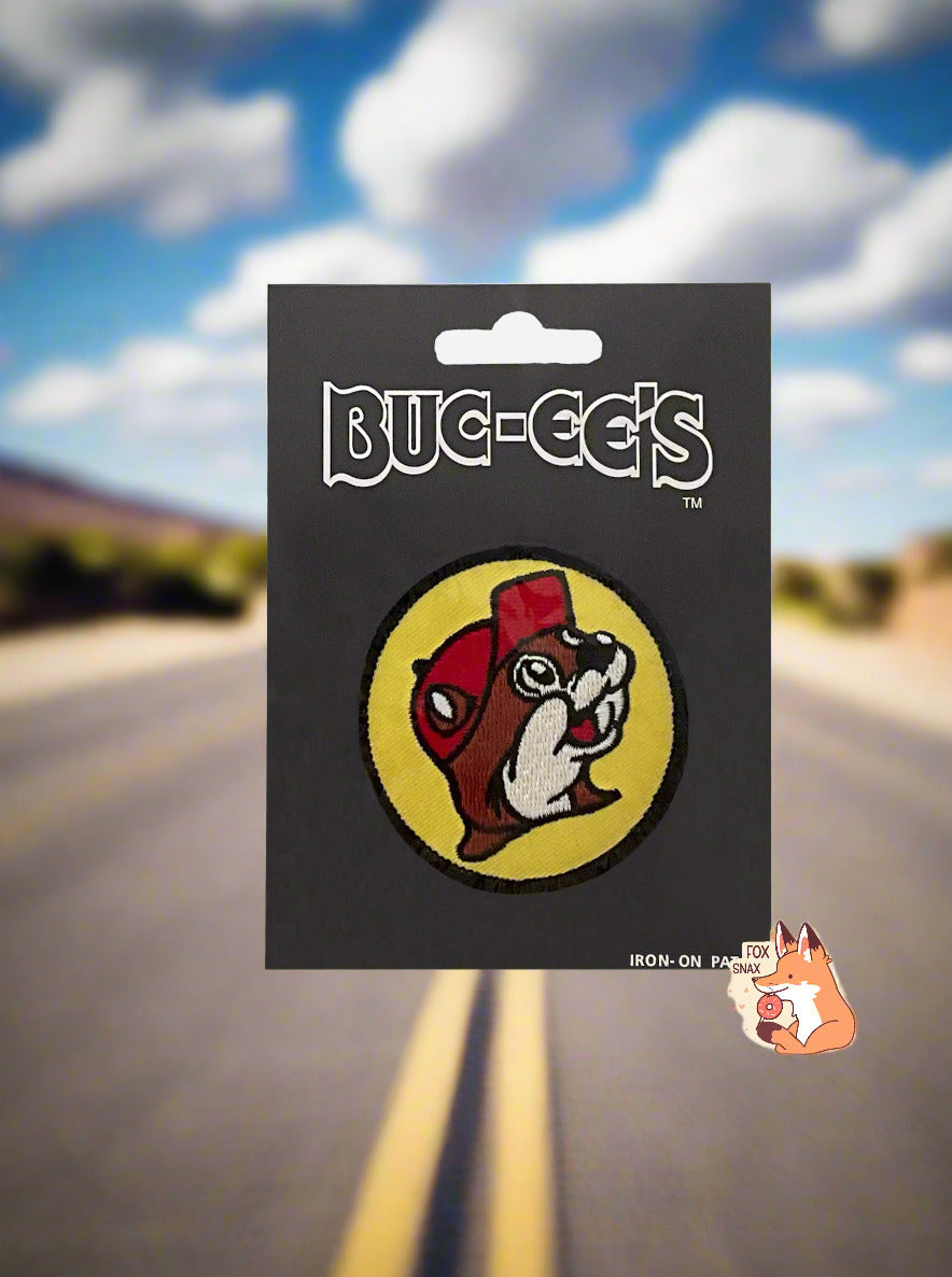 A picture of a circular iron-on patch. The patch is the Buc-ee's logo: a grinning beaver with a significant overbite and brown and white fur, wearing a red ballcap. Buc-ee is surrounded with a yellow circle, which is lined with black. The patch is hovering in midair above a pretty roadway; hills dotted with trees can be seen on the side, framed by a cloudy blue sky.