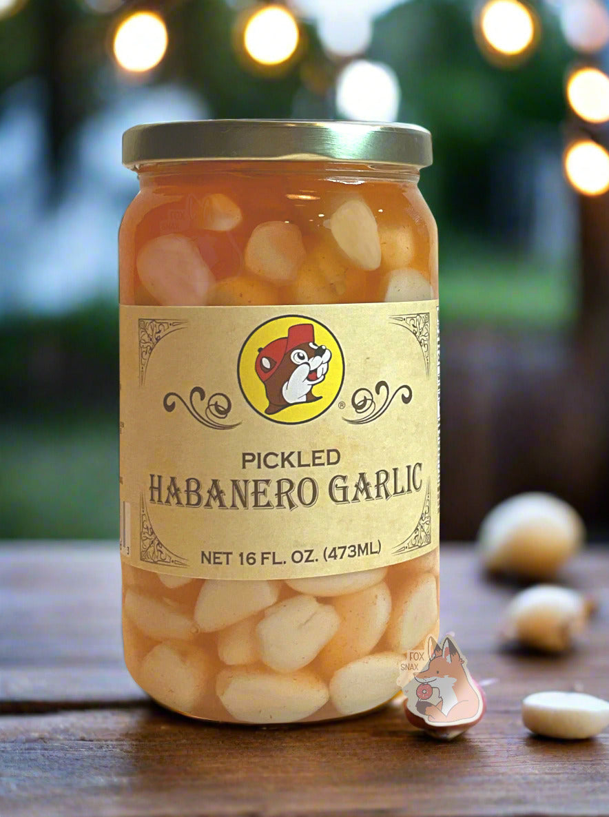 A picture of the front of a glass jar of Buc-ee's Pickled Habanero Garlic - the label is a "old-timey" sort of white/yellow with flared edges. The middle has a giant Buc-ee the Beaver logo on it. In the jar, floating garlic cloves in an orange habanero brine can be seen. The jar is sitting on a fancy dark wood outdoor table; garlic cloves litter the table. The background shows a bit of greenery - trees with string lights hanging off of them; clearly a party