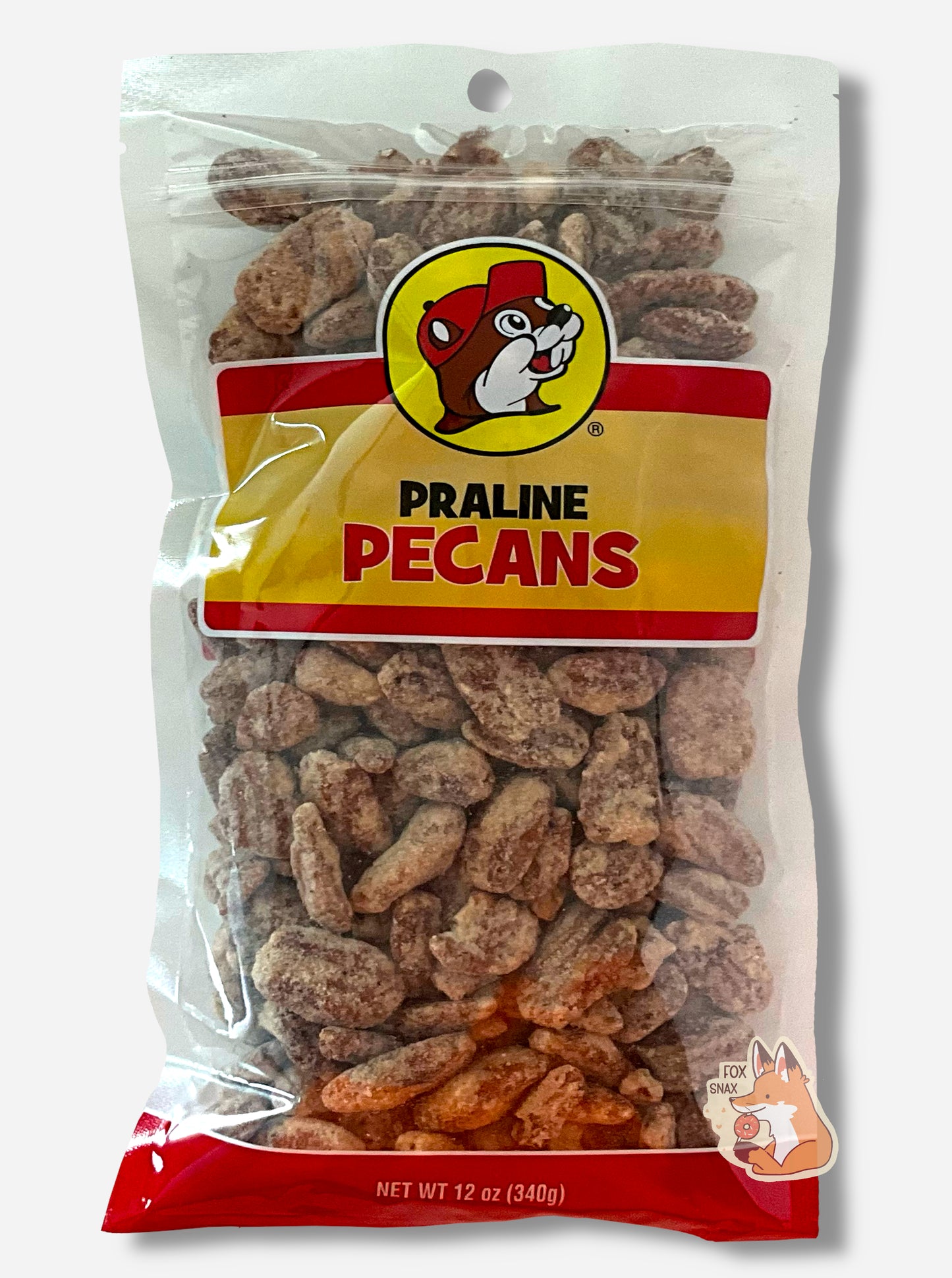 A picture of a clear plastic bag of Buc-ee's Praline Pecans. The Buc-ee's logo can be seen in the top center of the bag, then underneath that in black and red text, PRALINE PECANS. The bottom of the bag is red and reads Net Wt 12 oz. Inside the bag are a large number of dried pecans, covered in brownish sugary praline material.