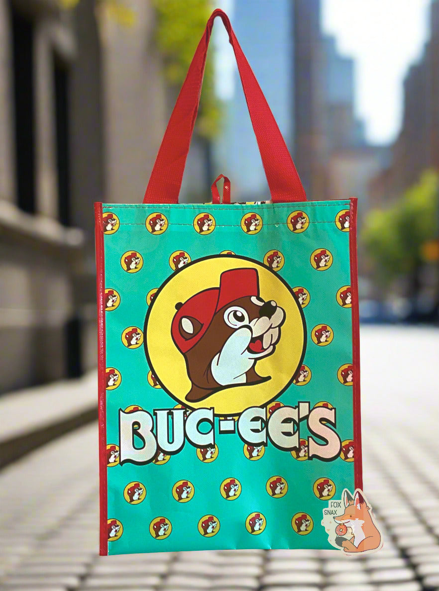 A picture of a large rectangular turquoise plastic shopping bag with multiple small Buc-ee The Beaver logos on it.  The bag itself has a large Buc-ee's logo right in the middle and the word BUC-EE'S in white text underneath of it.  The strap on the bag is made of red  cloth. The vertical edges of the bag have a thin red stripe.  The background shows a grey cobblestone-lined city street, framed with impressive buildings.