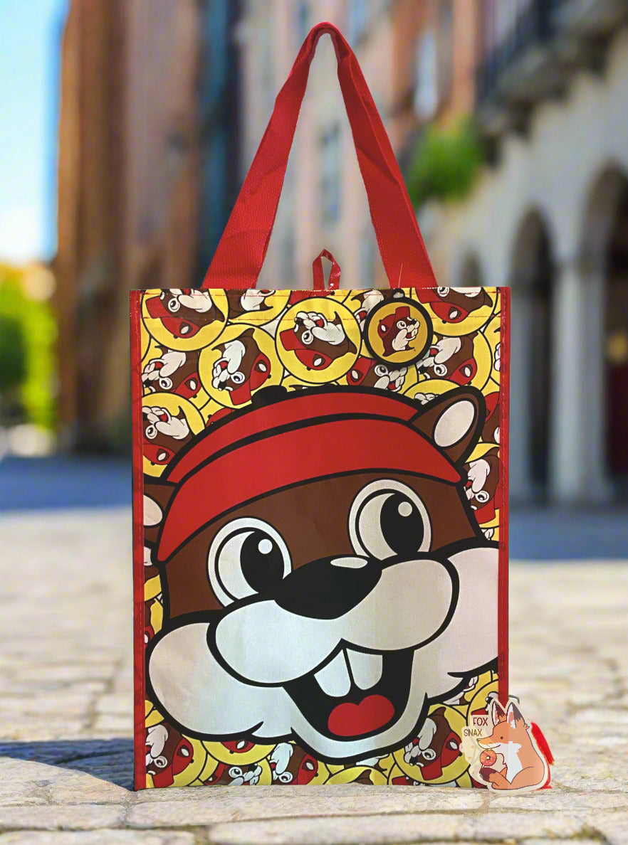 A picture of a large rectangular red plastic shopping bag with the multiple Buc-ee The Beaver logos on it.  The bag itself has a large closeup of Buc-ee's face right in the middle.   The strap on the bag is made of red  cloth.  The vertical edges of the bag have a thin red stripe.  The background shows a grey cobblestone-lined city street, framed with impressive buildings.